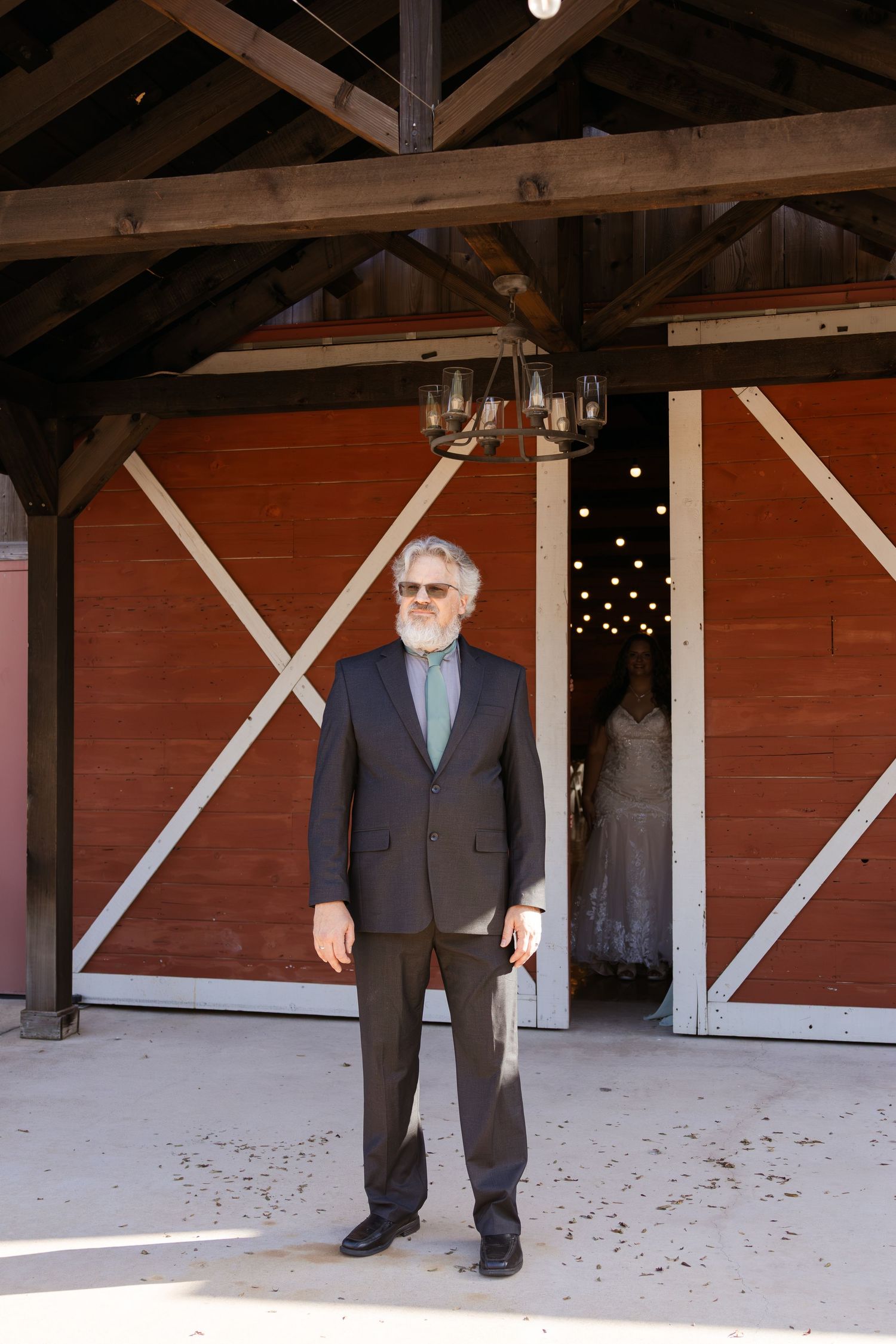 Older man in dark suit with gray beard stands before rustic red barn doors at an outdoor wedding venue.