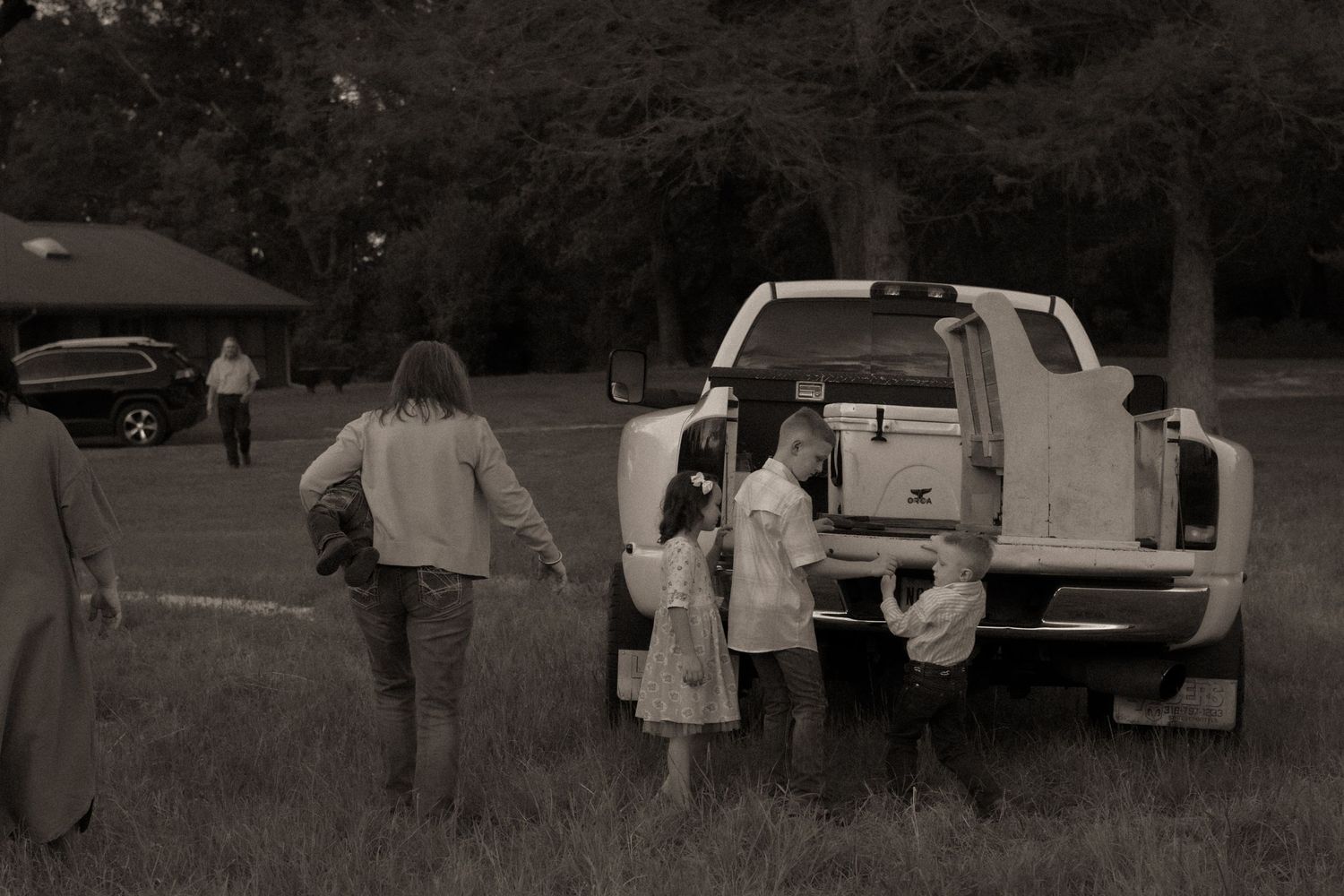 Black and white photo of children gathering around a pickup truck tailgate in a rural field with trees.