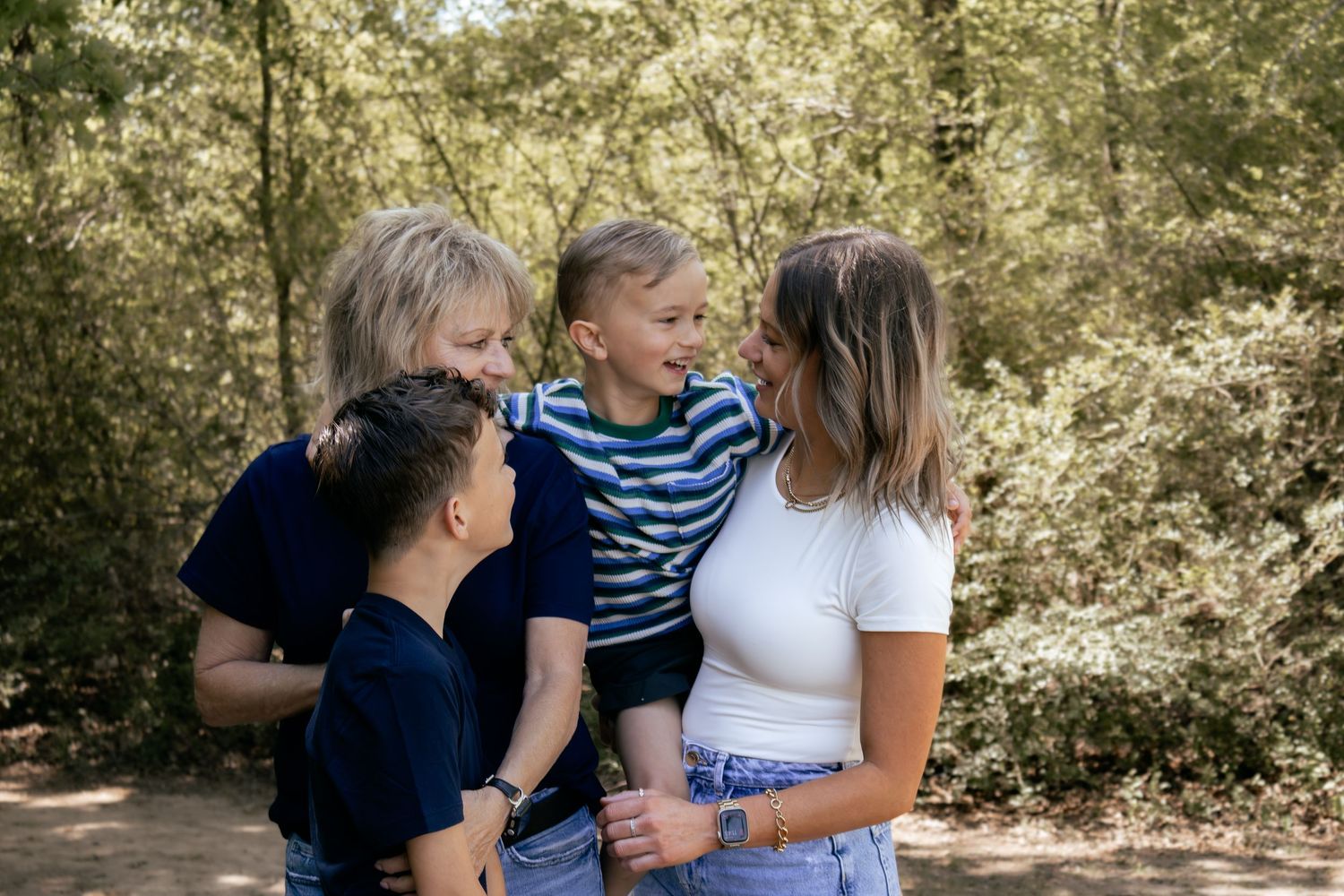Family of four smiling and laughing together on a sunny forest path, two adults holding young children outdoors.