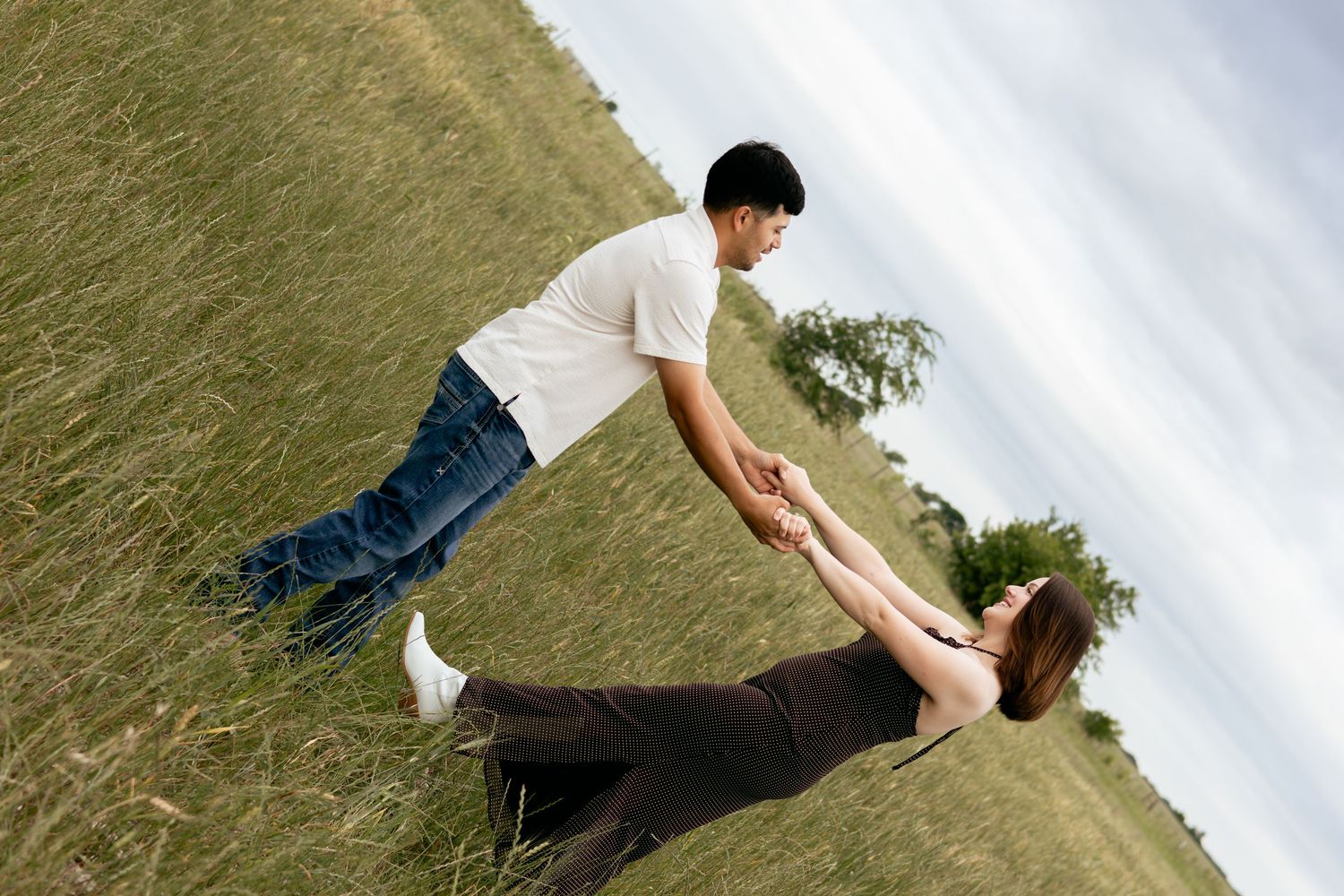 Couple holding hands in a grassy field, man in white shirt leaning back while woman in black dress dips gracefully.