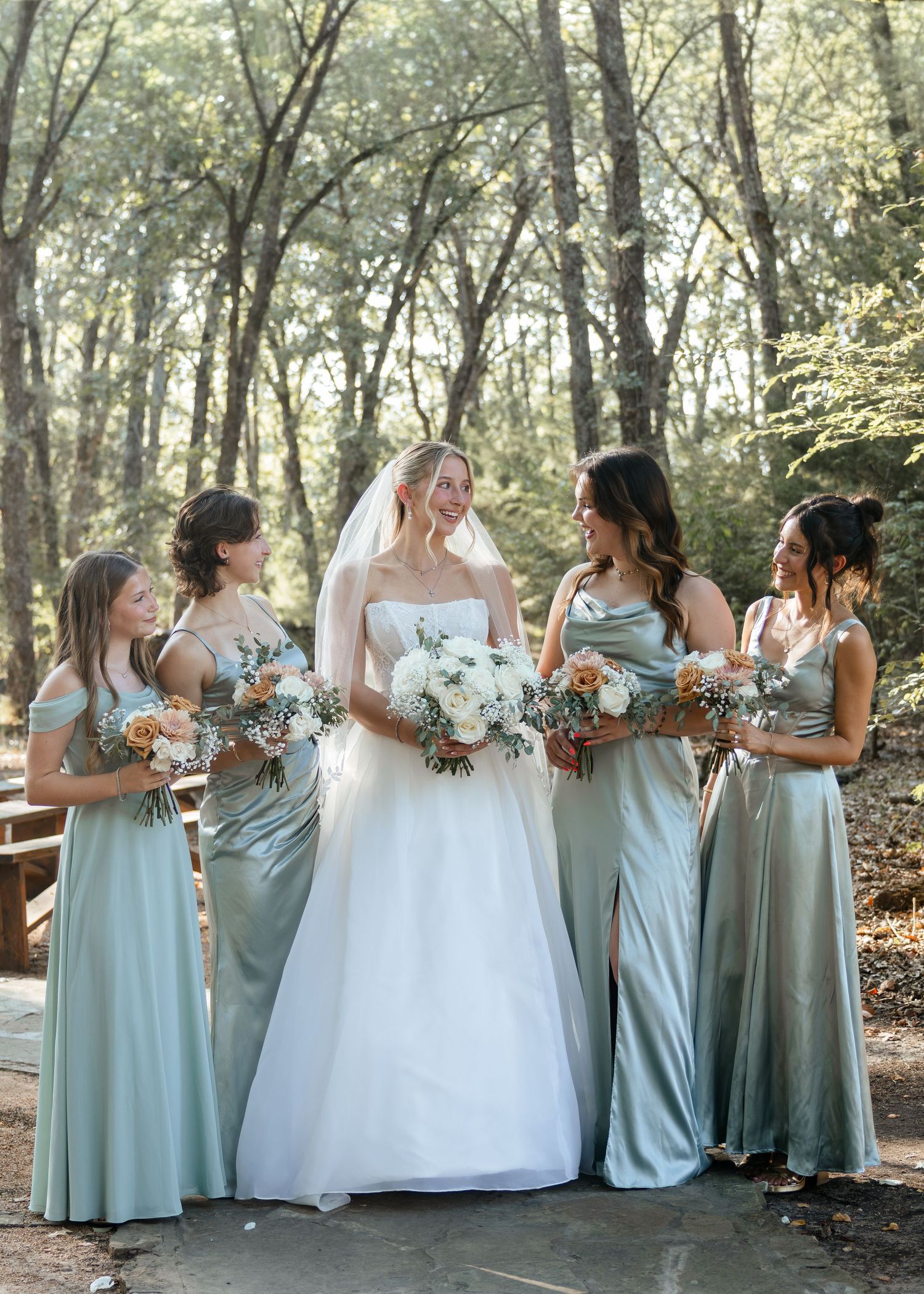 Bride in white gown with four bridesmaids in sage green satin dresses holding bouquets in a wooded outdoor setting.