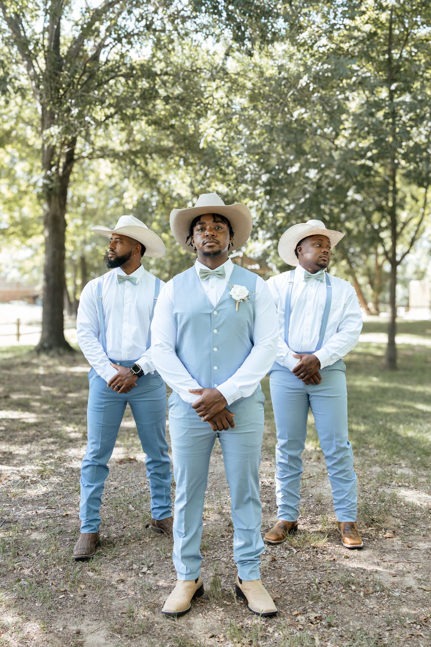 Groom and groomsmen in light blue suits, suspenders, and cowboy hats posing outdoors under shaded trees.