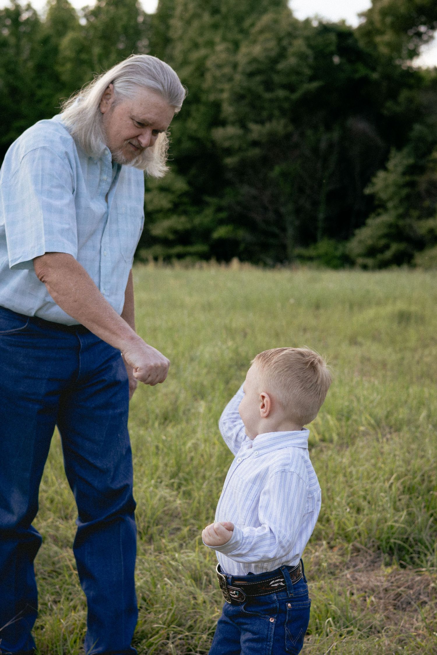 Elderly man with long gray hair bending down to interact with a young boy in a green field.