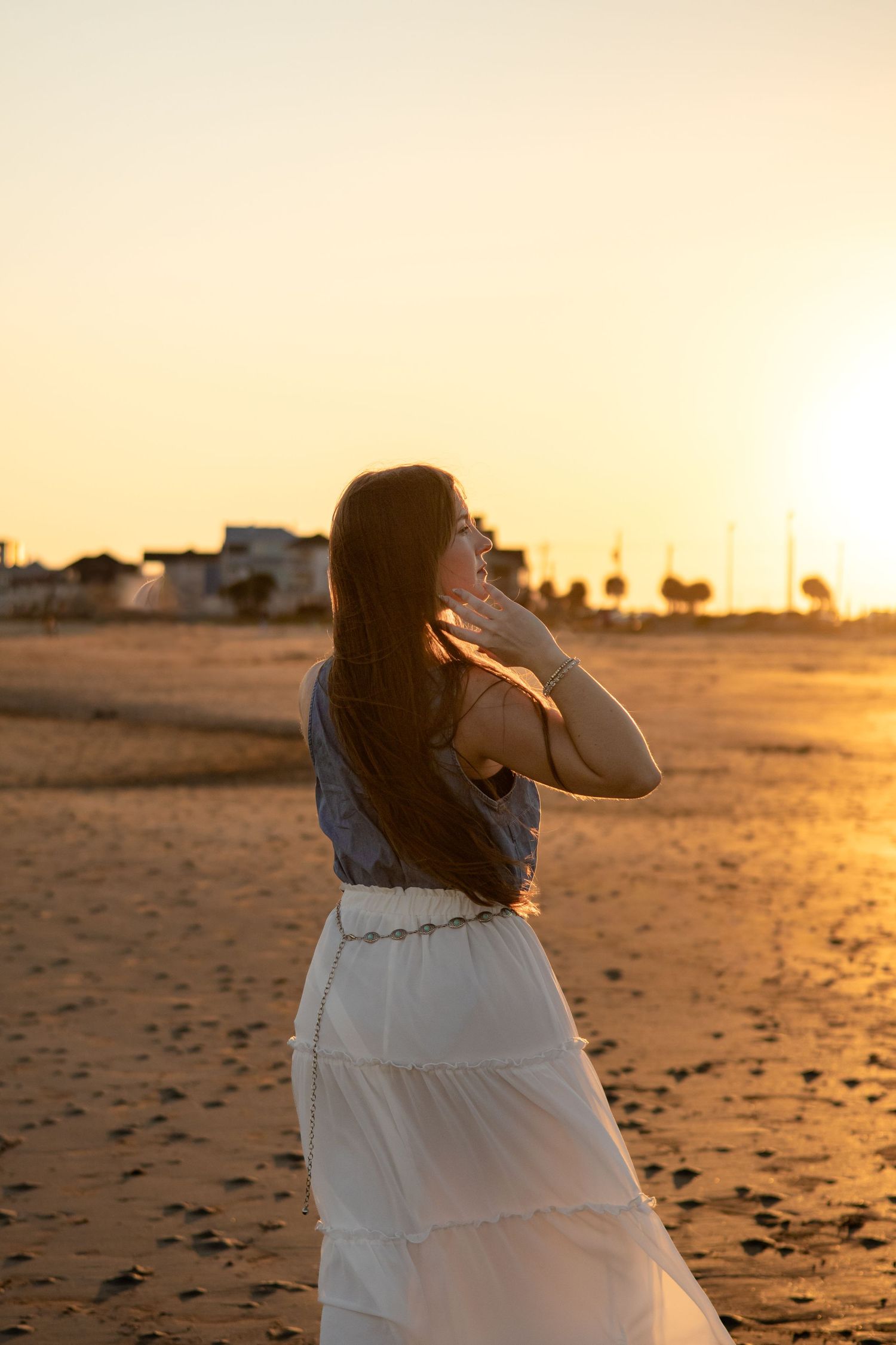 Woman in flowing white dress twirling on beach at sunset creating silhouette against golden sky.