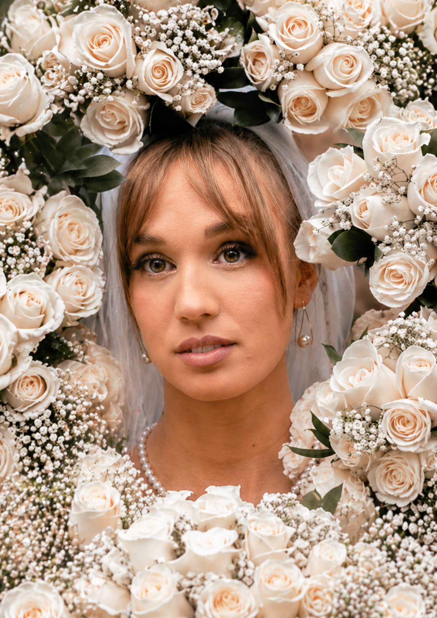 Bride framed by white roses and baby's breath, wearing a veil and pearl necklace, with a soft natural makeup look.