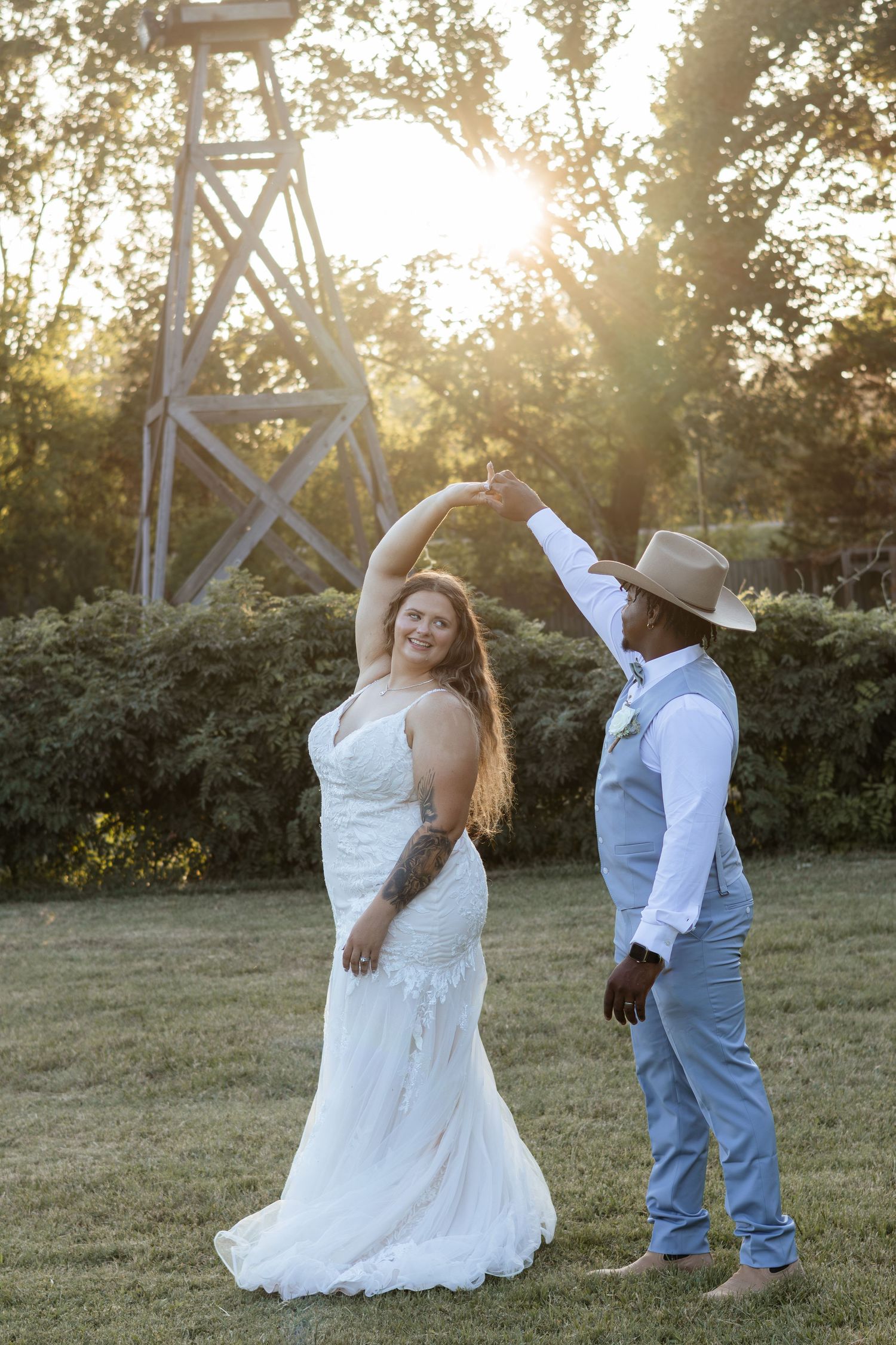 Bride and groom dancing at golden hour sunset outdoors, groom in cowboy hat twirling bride in lace wedding gown.