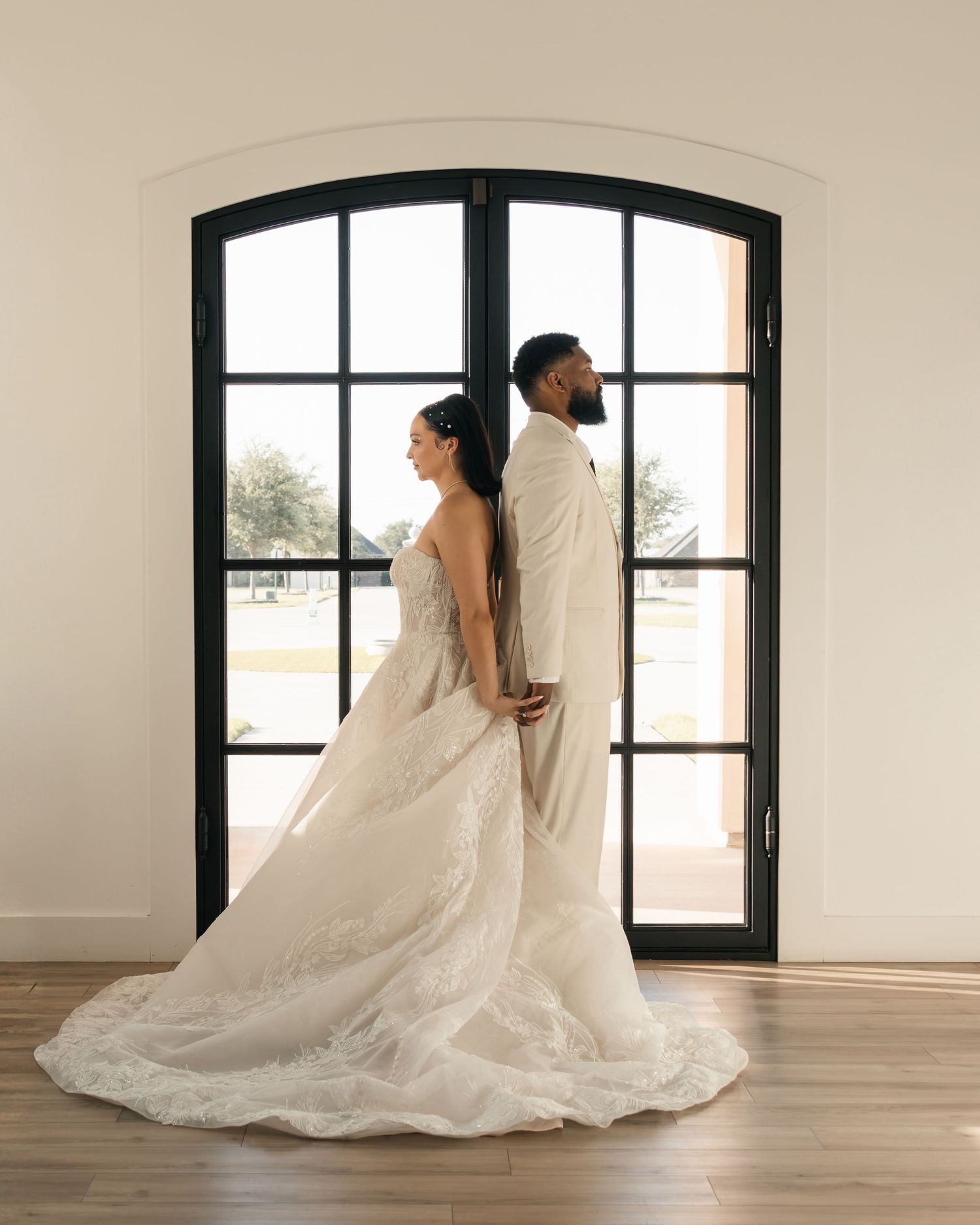 Bride in white lace gown and groom in ivory suit stand back-to-back before arched French doors on wedding day.