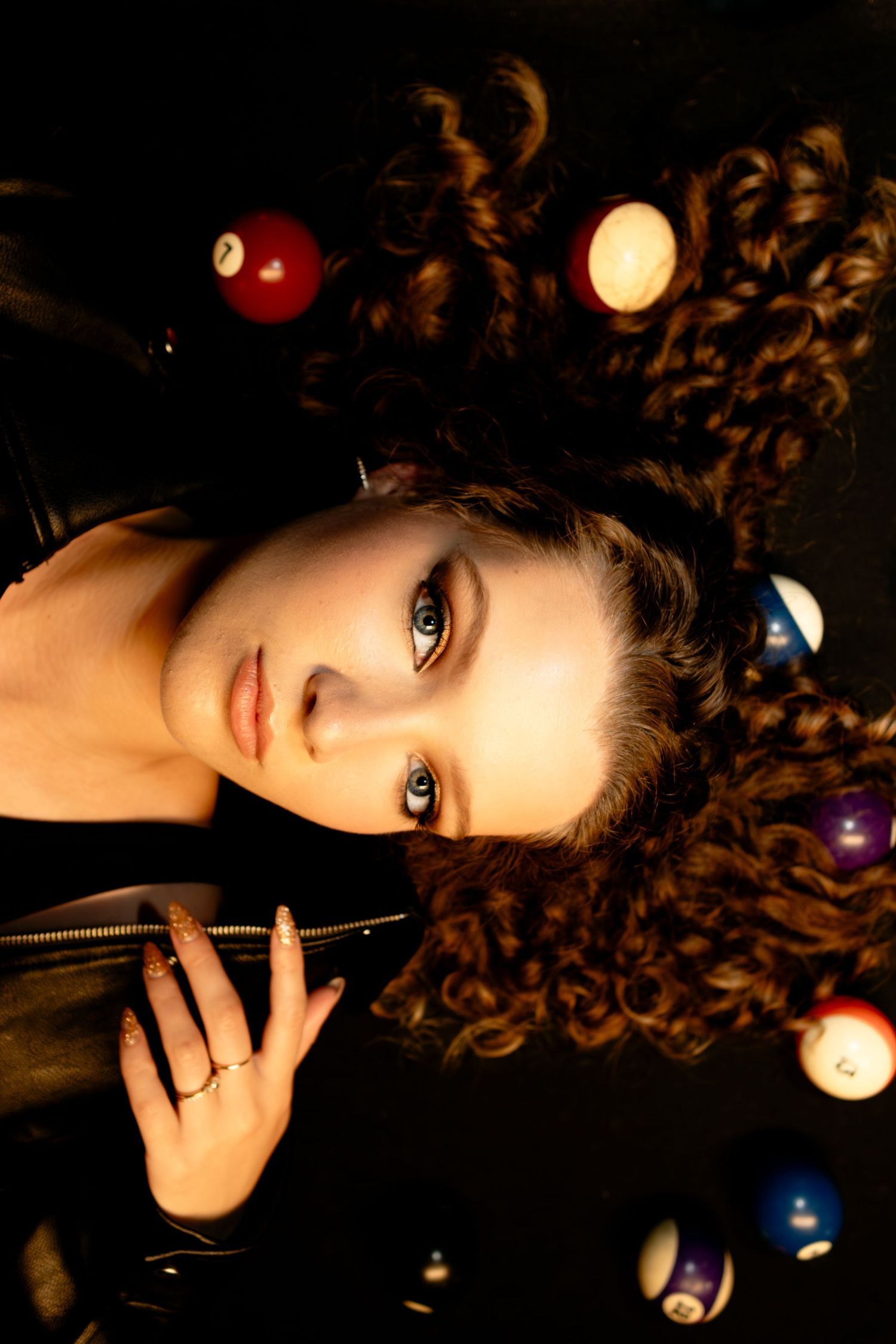 Close-up portrait of a curly-haired woman lying on a pool table surrounded by billiard balls.