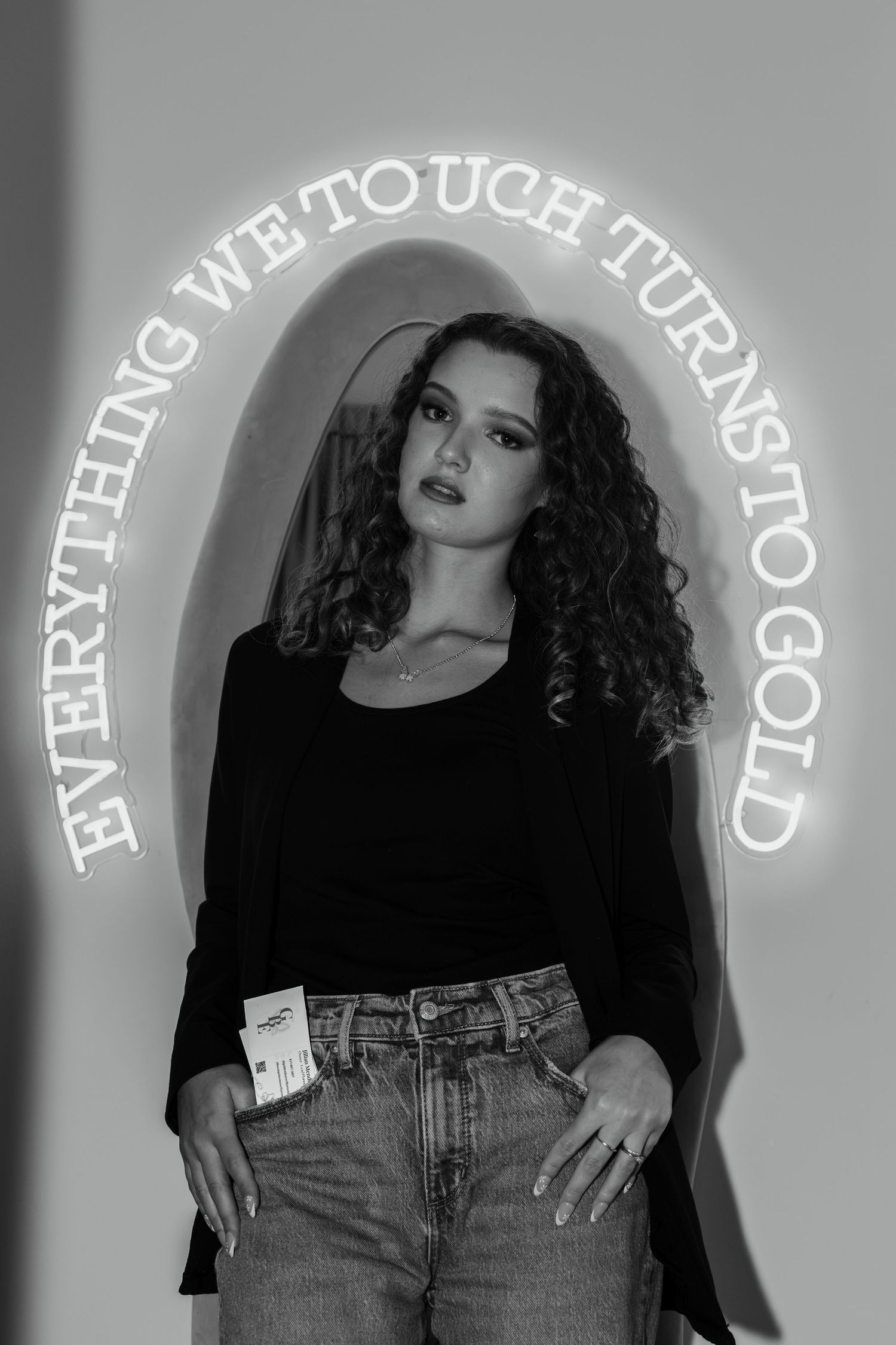 Black and white portrait of curly-haired woman in black outfit with neon gold quote sign behind her.