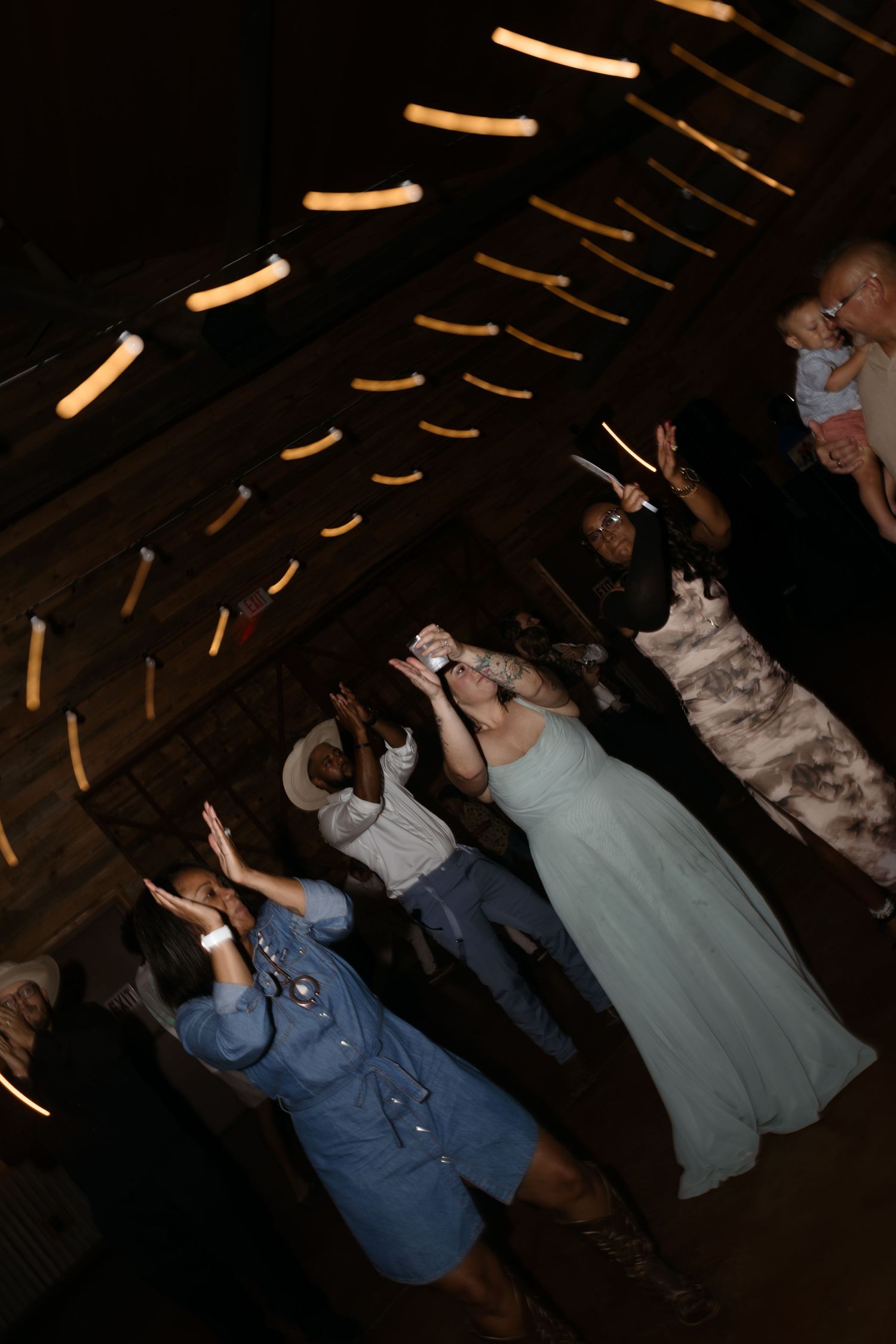 Wedding reception guests dancing under warm string lights inside a rustic barn venue during evening celebration.