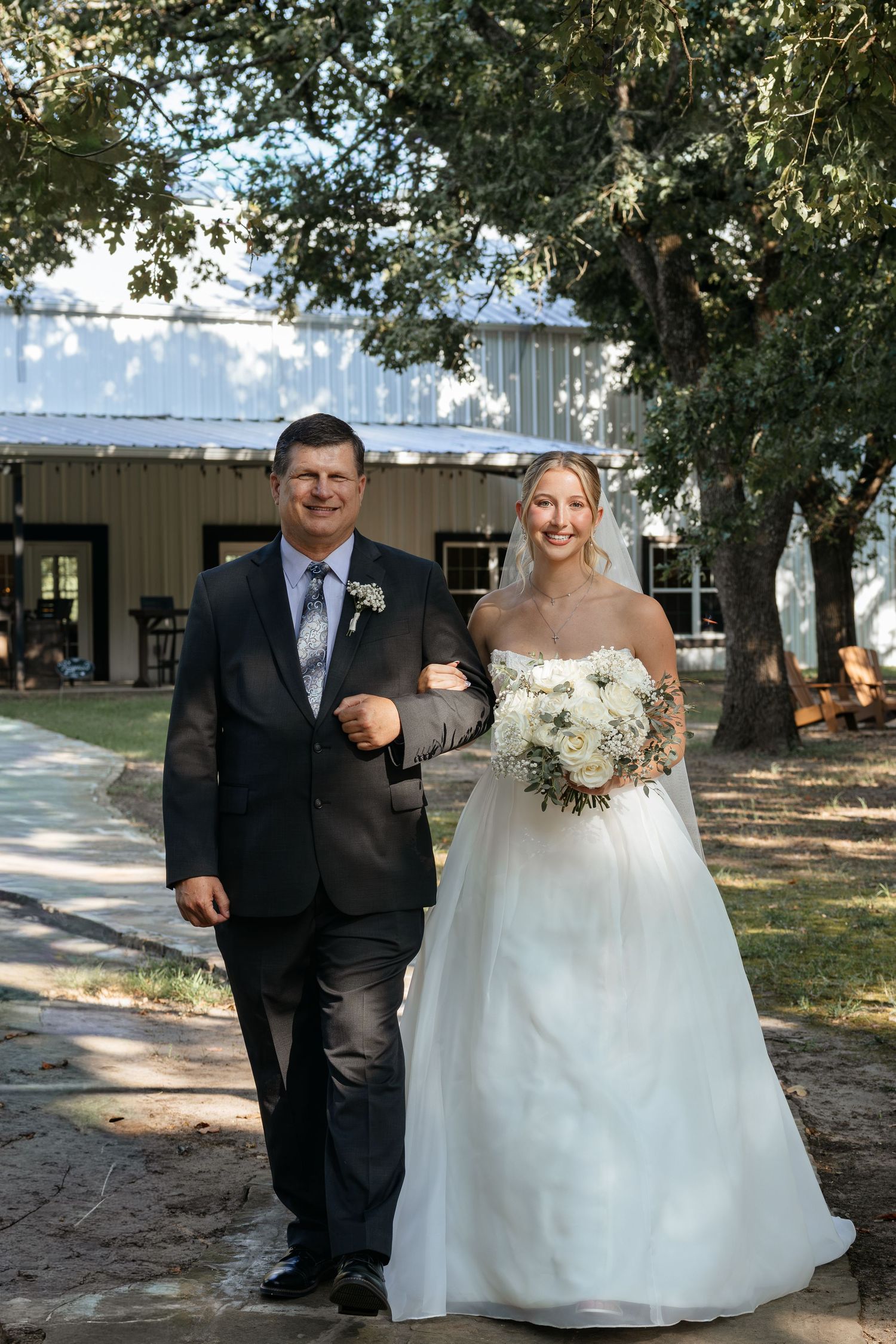 Bride in white strapless gown holding bouquet walks arm-in-arm with father in dark suit toward wedding ceremony.