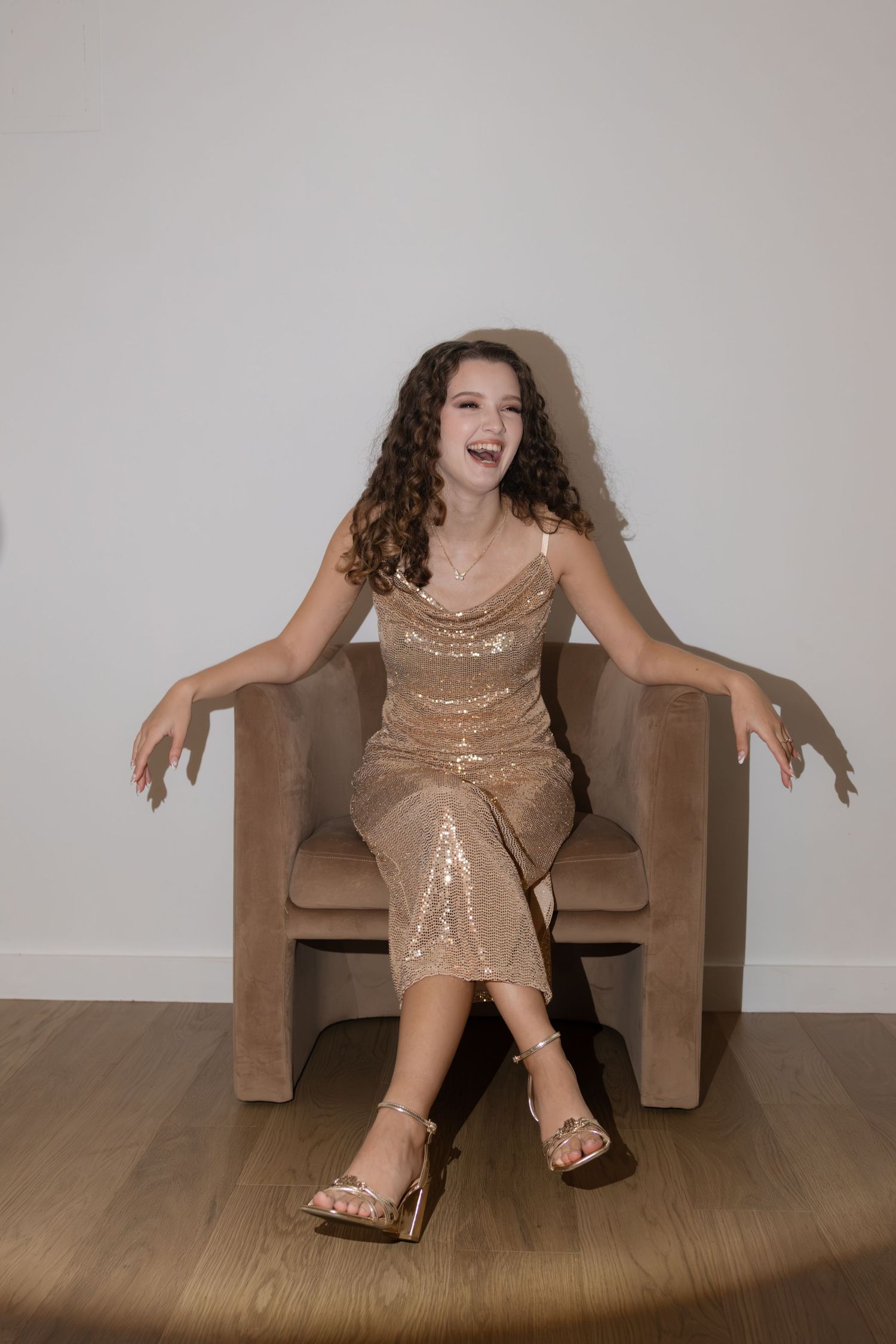Laughing young woman in gold sequin midi dress seated in beige velvet chair on wooden floor.