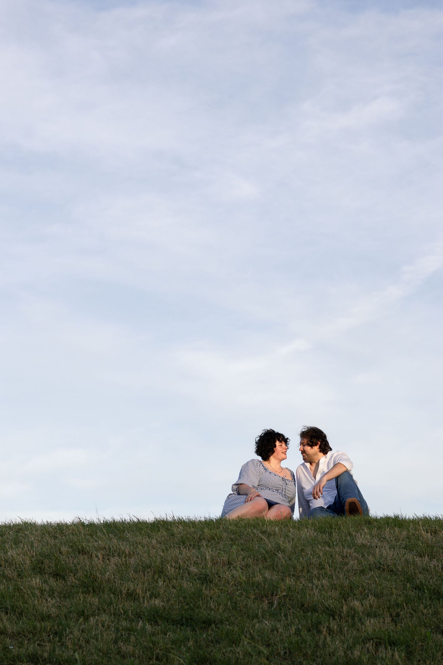 Romantic couple sitting together on a grassy hill, gazing at each other under a cloudy blue sky.