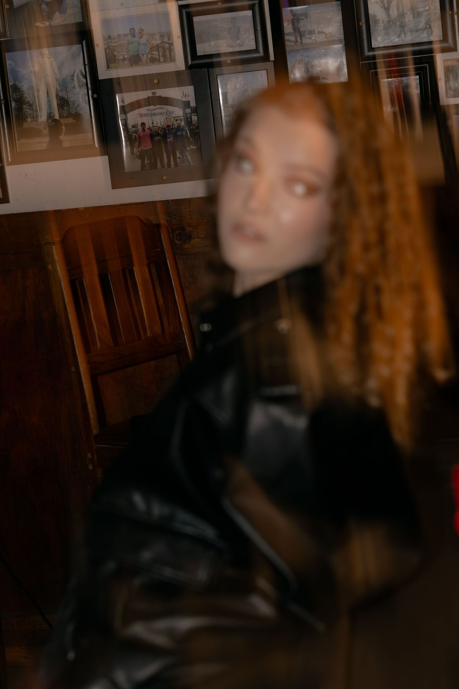 Artistic blurred portrait of curly-haired woman in black leather jacket against wall of framed photos.