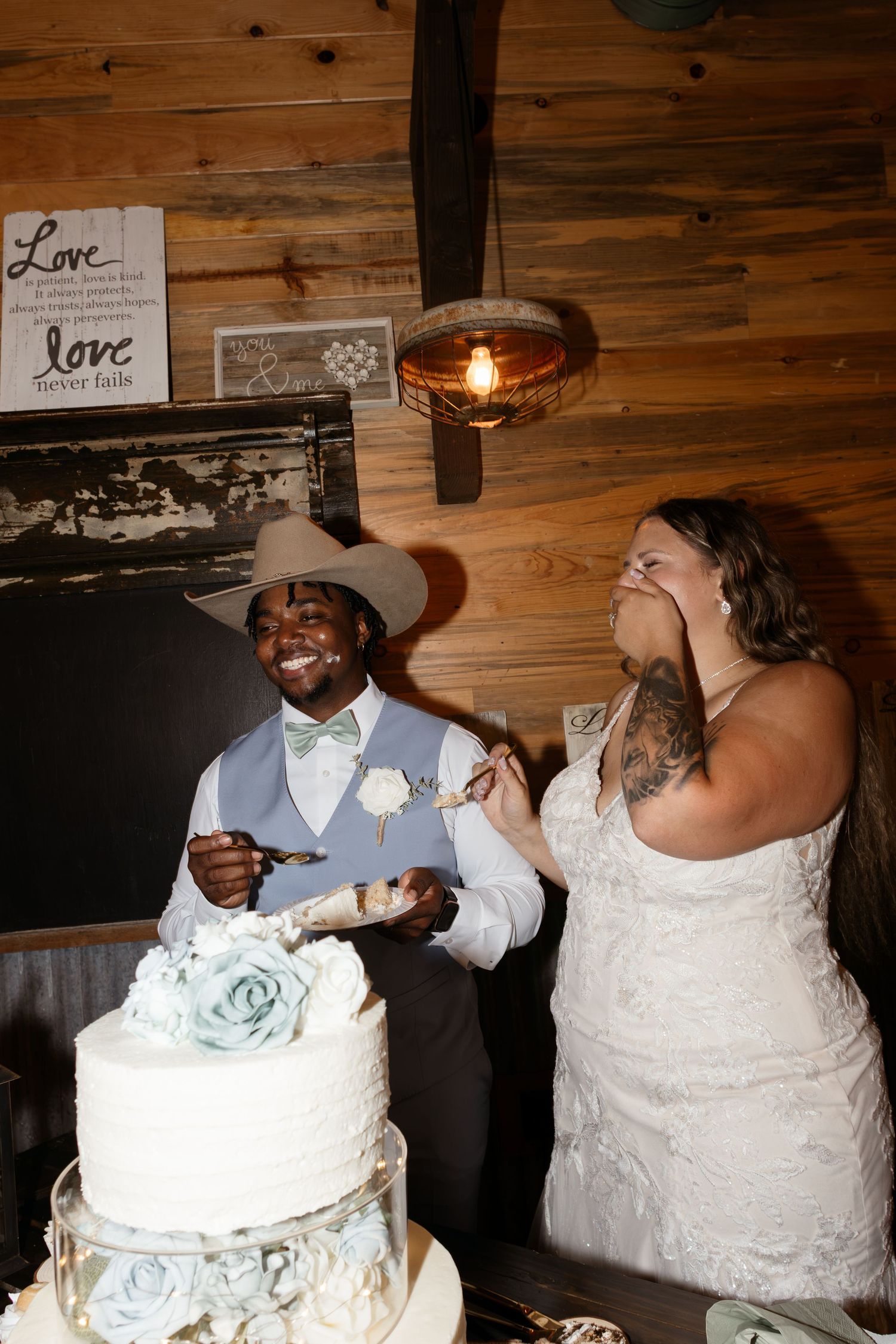 Smiling bride and groom cutting white wedding cake decorated with blue roses in rustic barn reception venue.