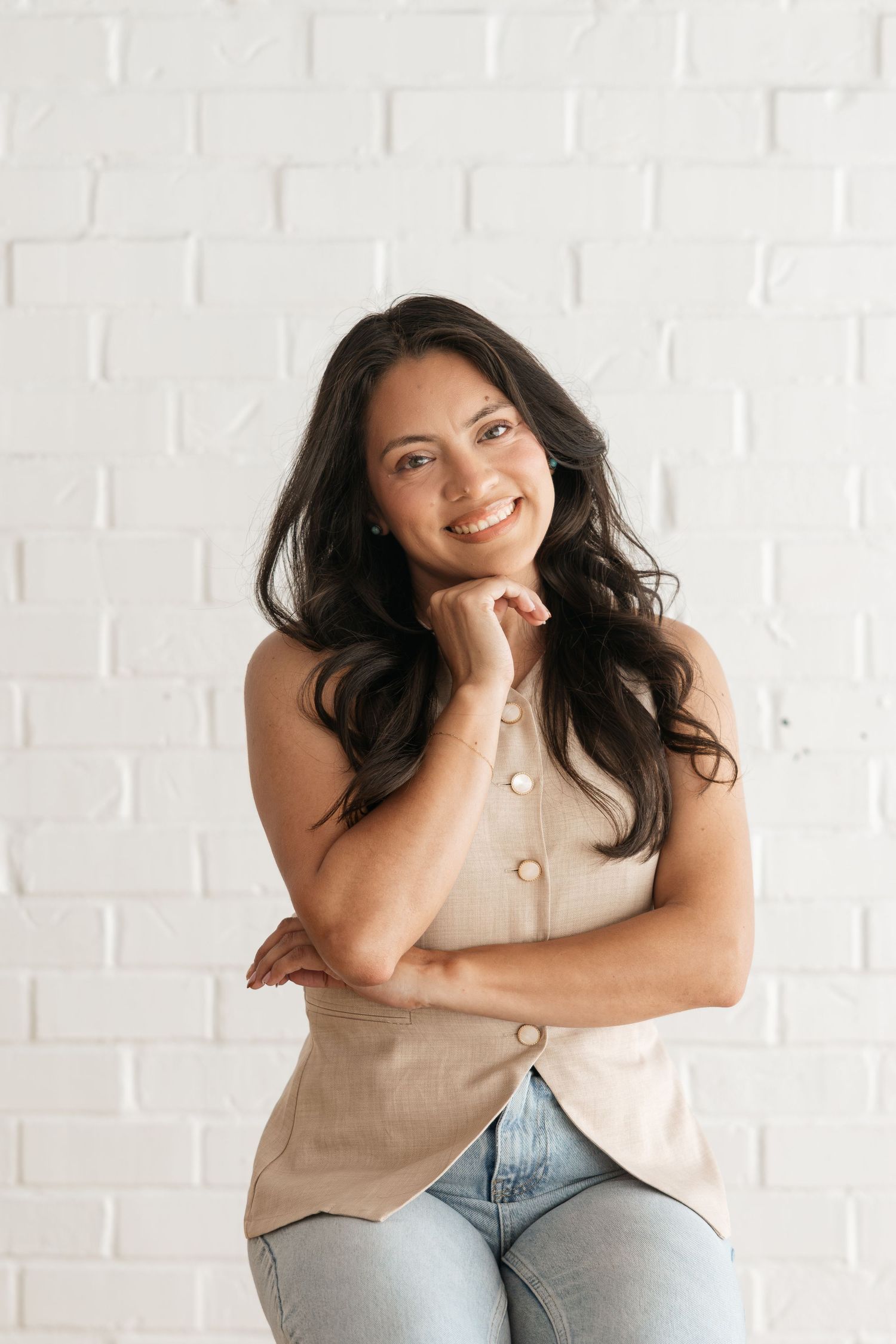 Smiling woman with dark wavy hair wearing a beige sleeveless top and jeans, posing against a white brick wall.