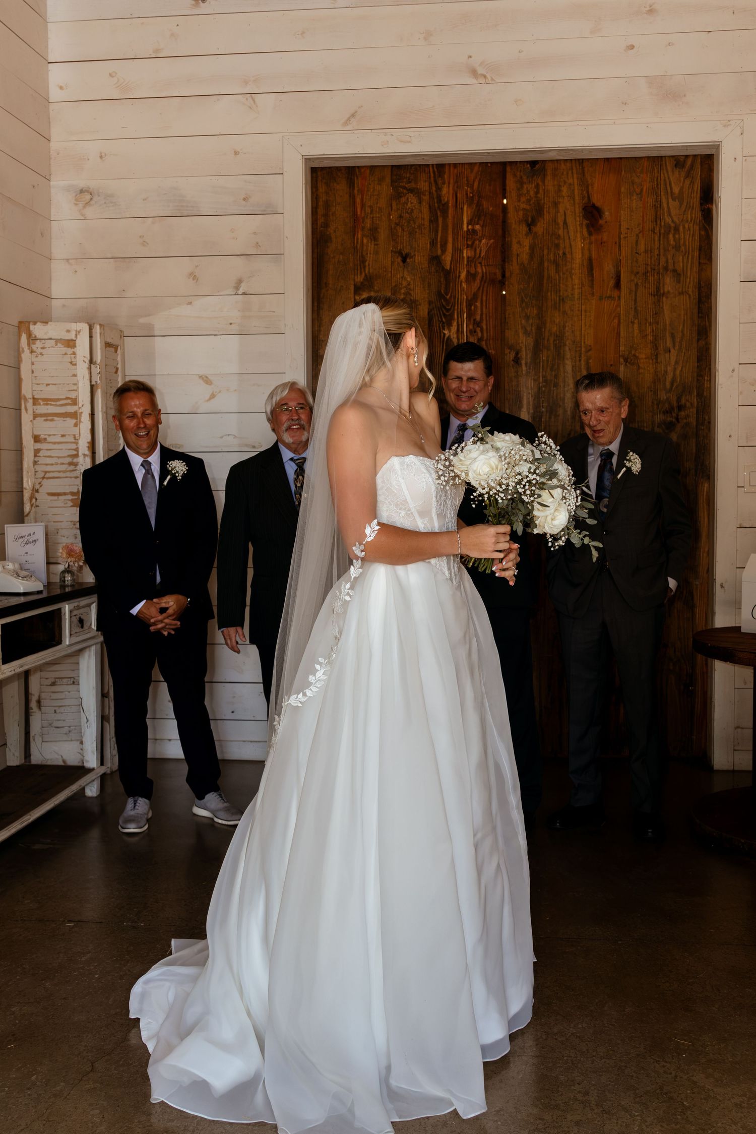 Bride in white gown and veil holds bouquet before large wooden doors as smiling guests look on.