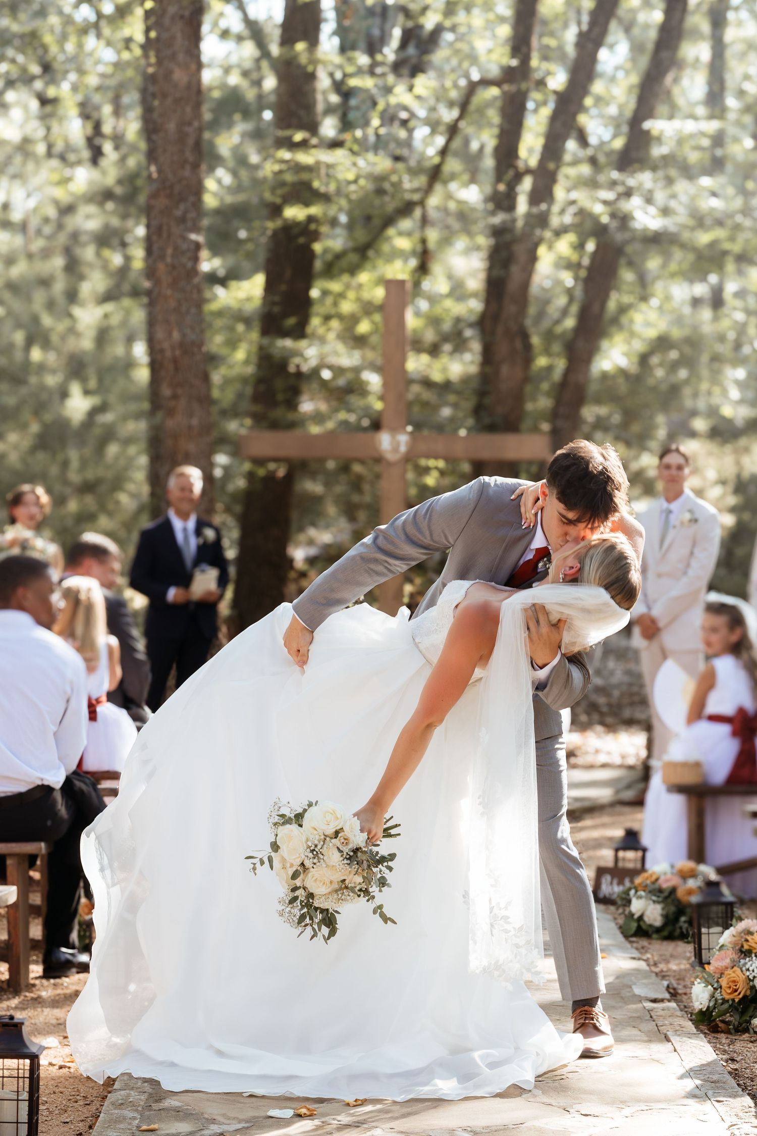 Groom dips bride for a romantic kiss at outdoor woodland wedding ceremony with wooden cross backdrop.