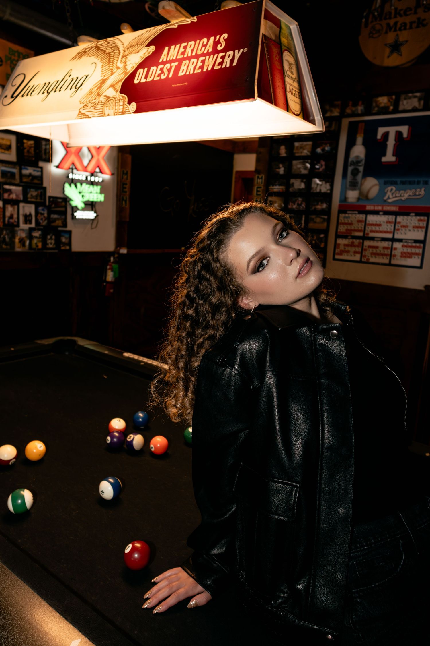 Young woman in black leather jacket poses by a pool table under a Yuengling brewery sign in a bar.
