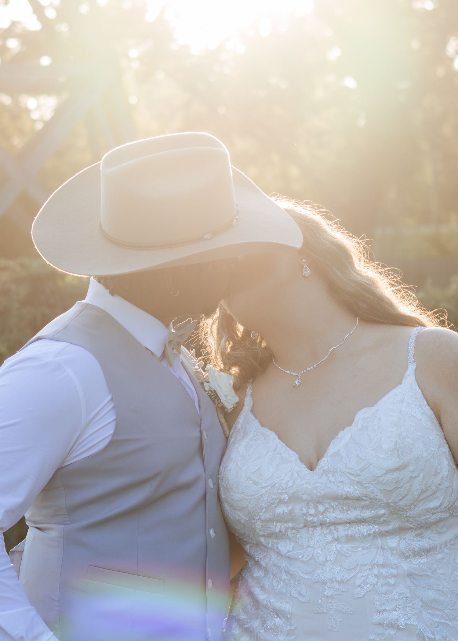 A bride and groom share a romantic kiss at sunset, with golden backlight glowing behind them in an outdoor wedding setting.