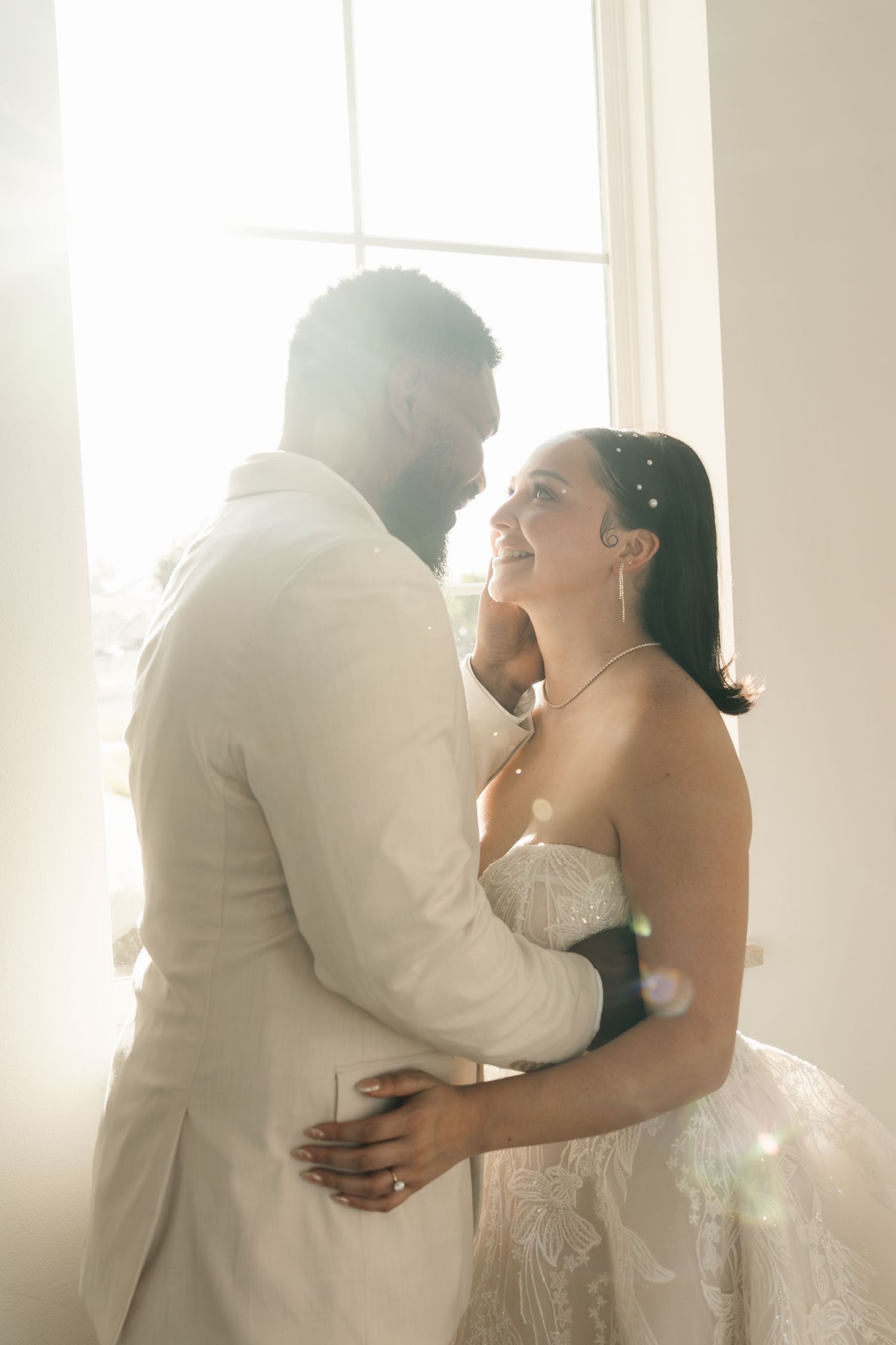 Bride and groom embrace near sunlit window, she smiles up at him in lace gown, he wears white suit.
