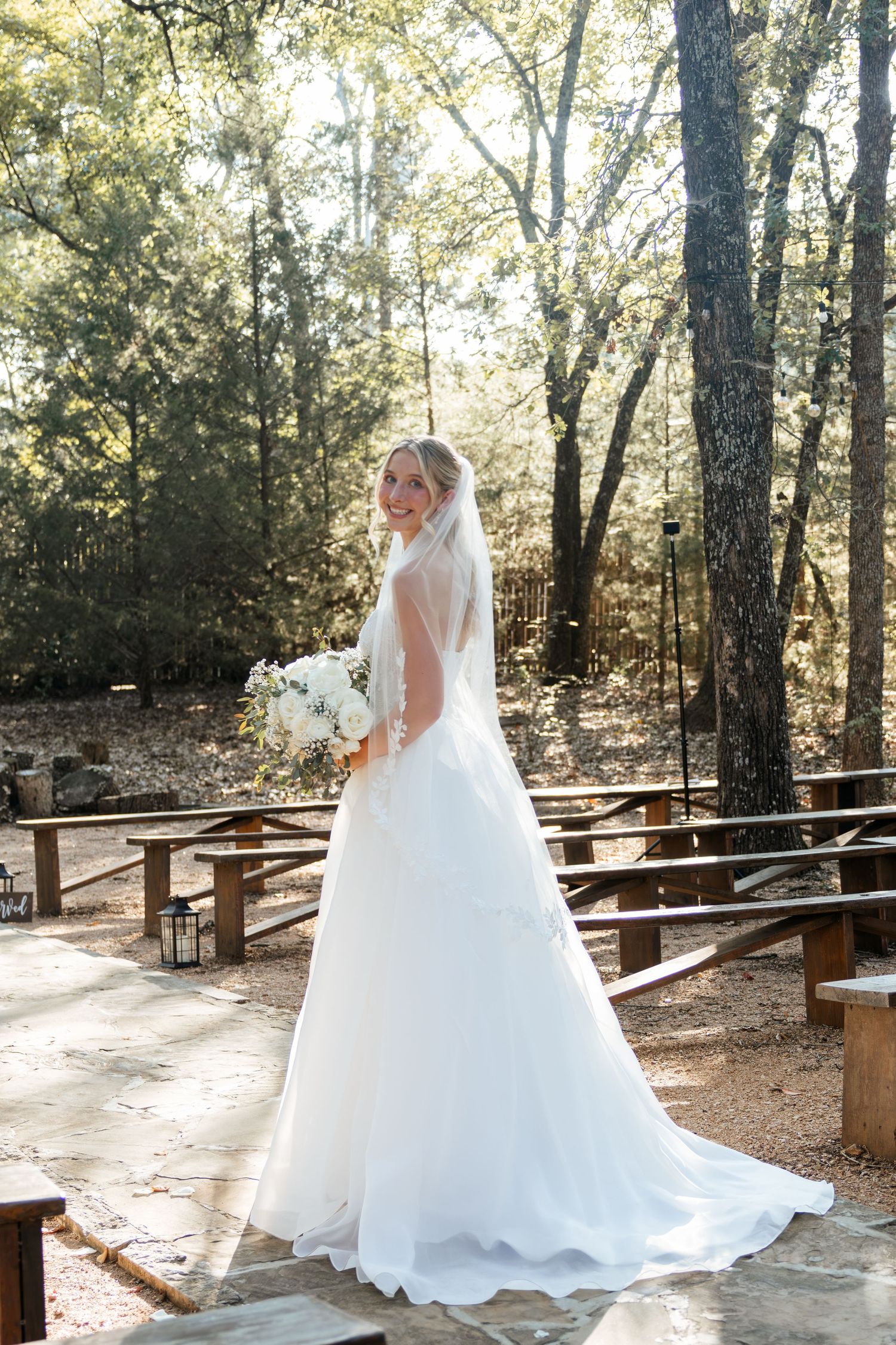 Radiant bride in flowing white ball gown and veil smiles while holding bouquet in outdoor woodland venue.