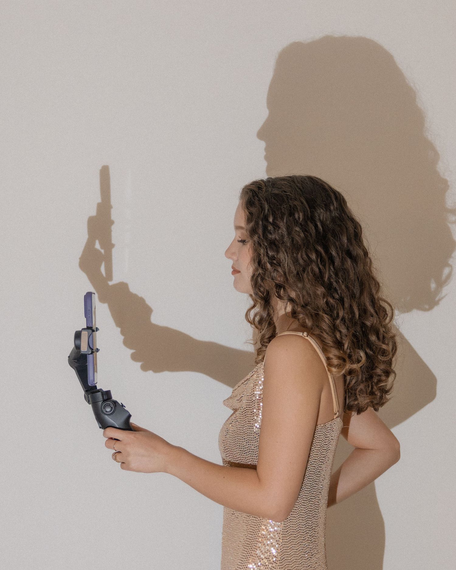Young woman in sequin dress holding a gimbal with dramatic shadow on white wall behind her.