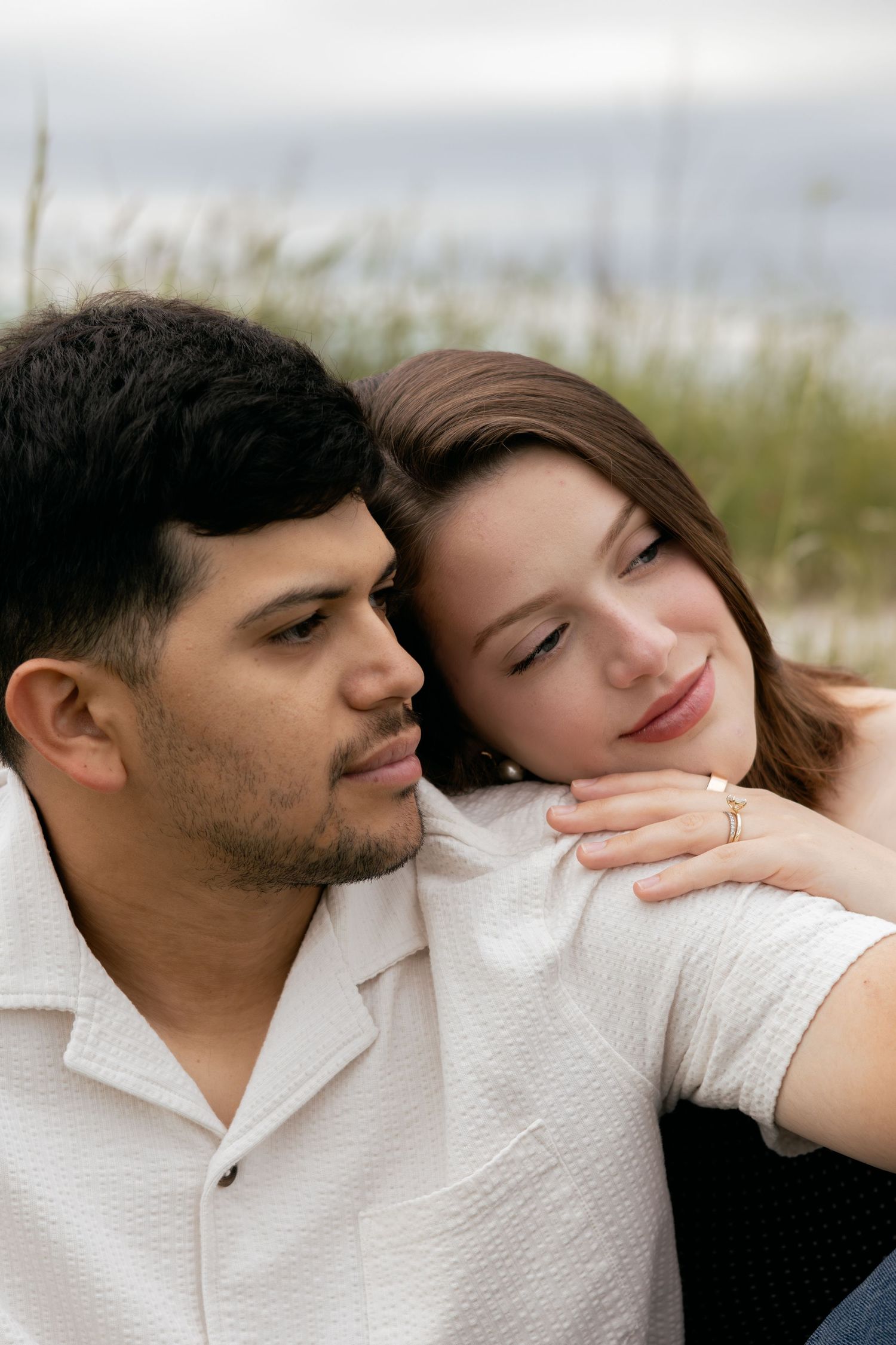 Couple sitting together near beach grass, woman resting head on mans shoulder tenderly.
