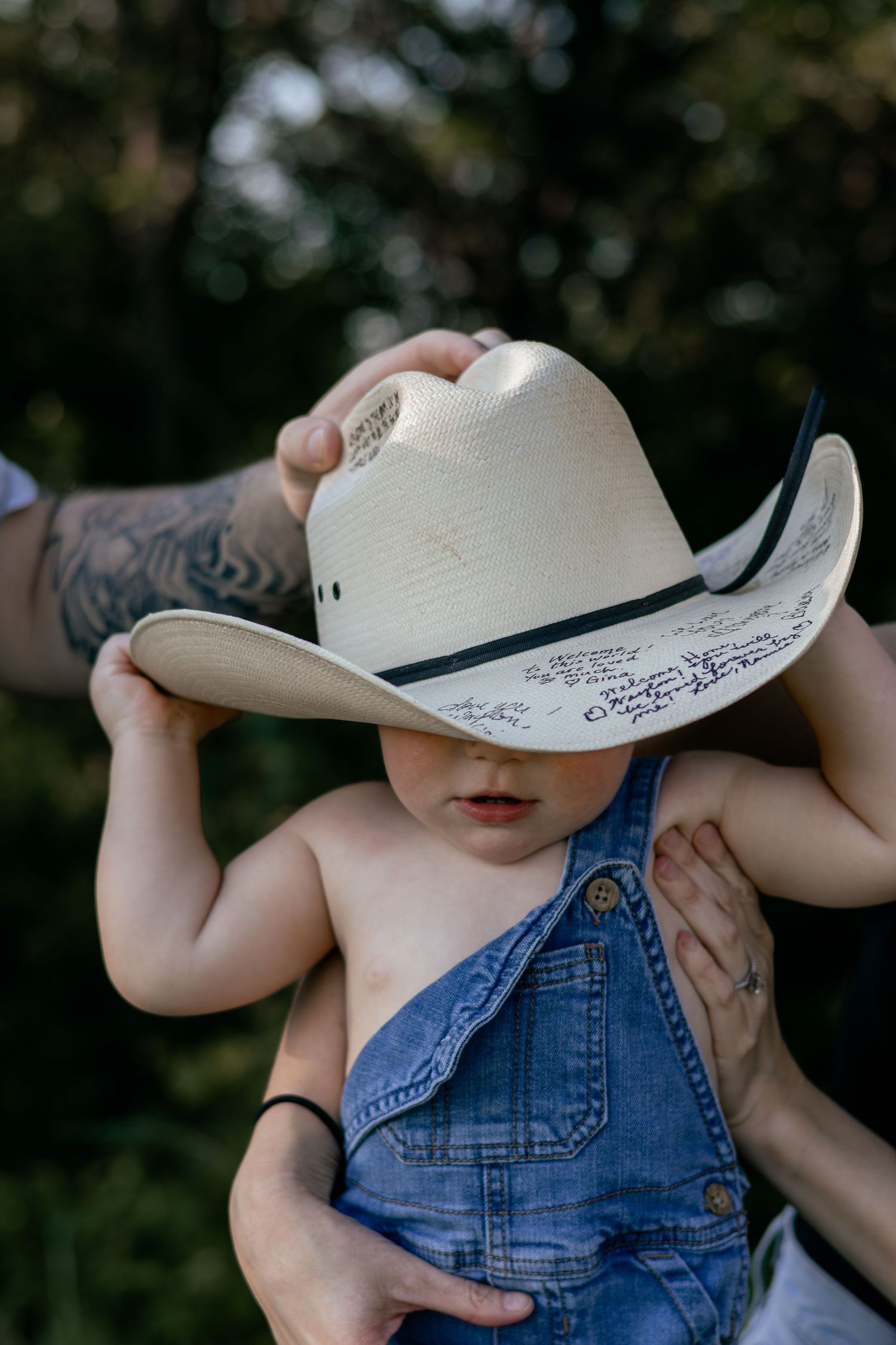 Baby in denim overalls wearing a large white cowboy hat held by tattooed adult arms outdoors.