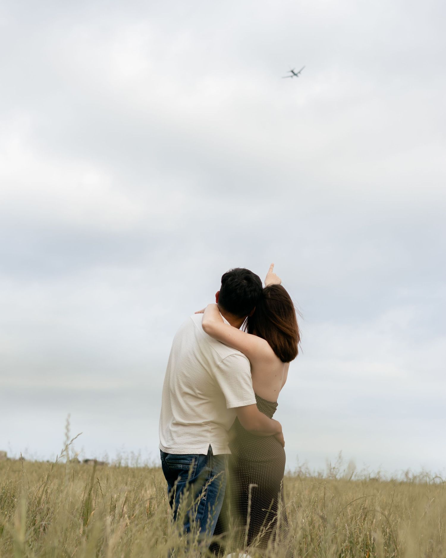 Couple embracing in a grassy field, pointing at a plane flying overhead under a cloudy sky.