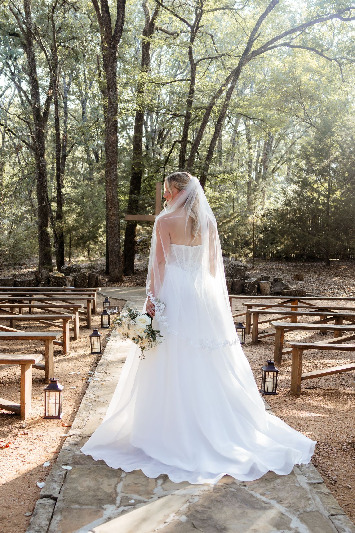 Bride in white gown and veil stands at outdoor woodland wedding aisle holding bouquet, surrounded by wooden pews and lanterns.