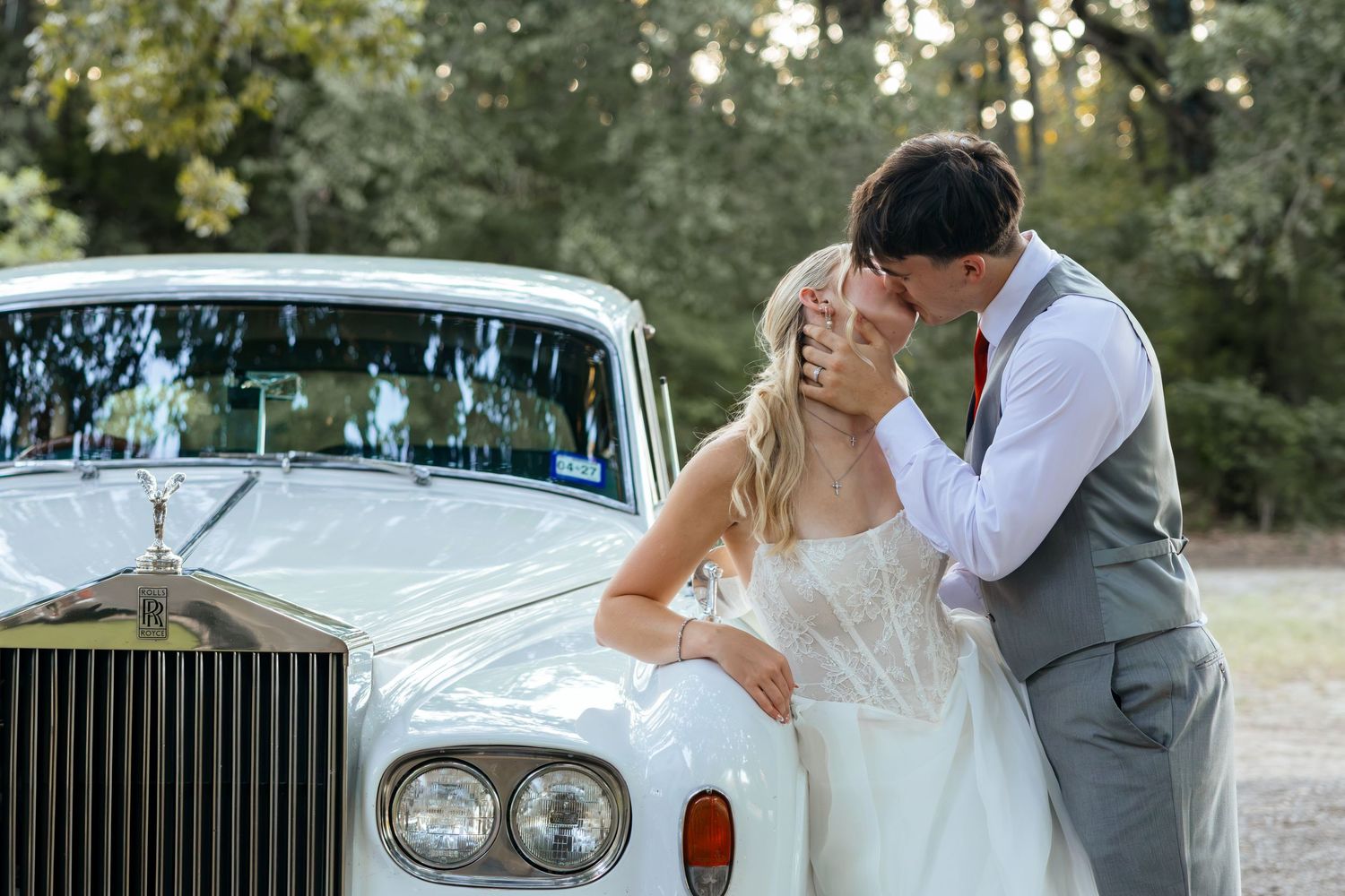 Bride and groom share a romantic kiss in front of a classic white Rolls-Royce at an outdoor wedding venue.