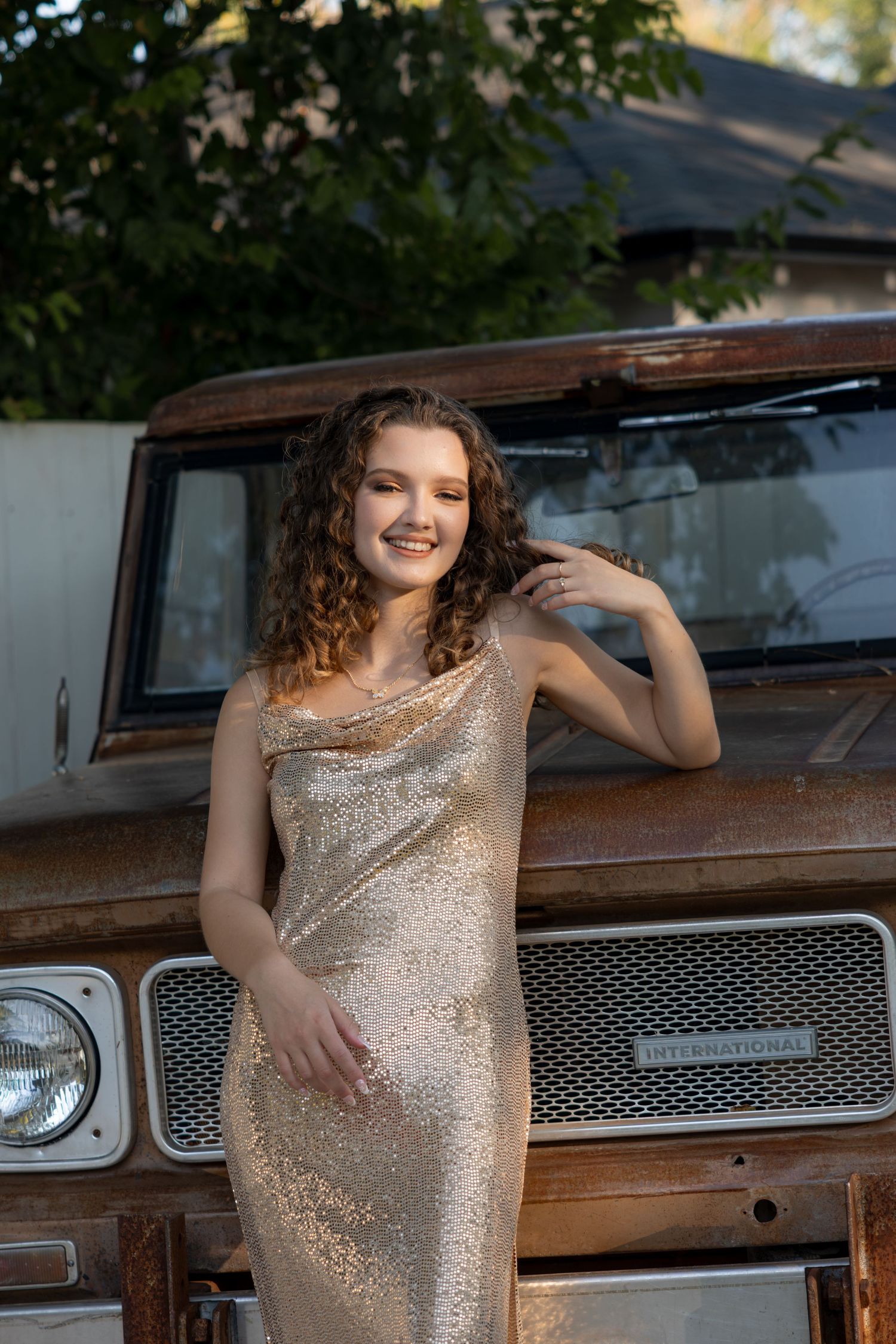 Smiling young woman in a gold sequin dress leans against a rusty vintage truck in a sunny outdoor setting.