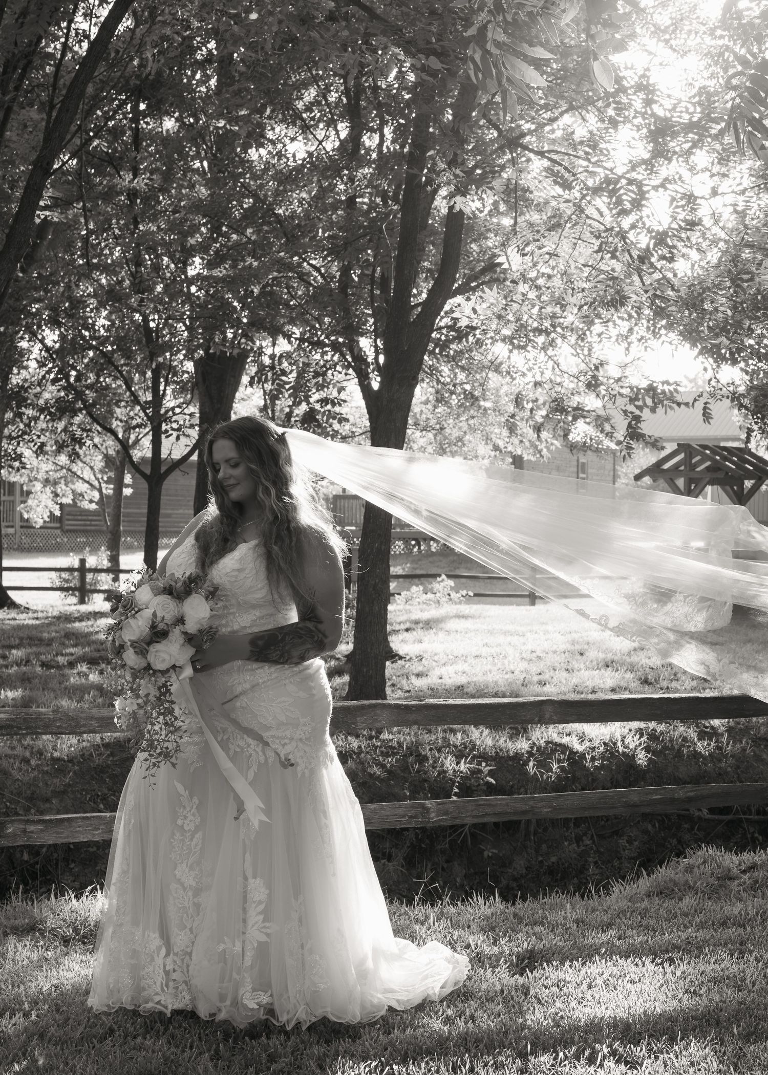 Black and white bridal portrait of a bride in lace gown with flowing veil caught in wind, holding bouquet outdoors.
