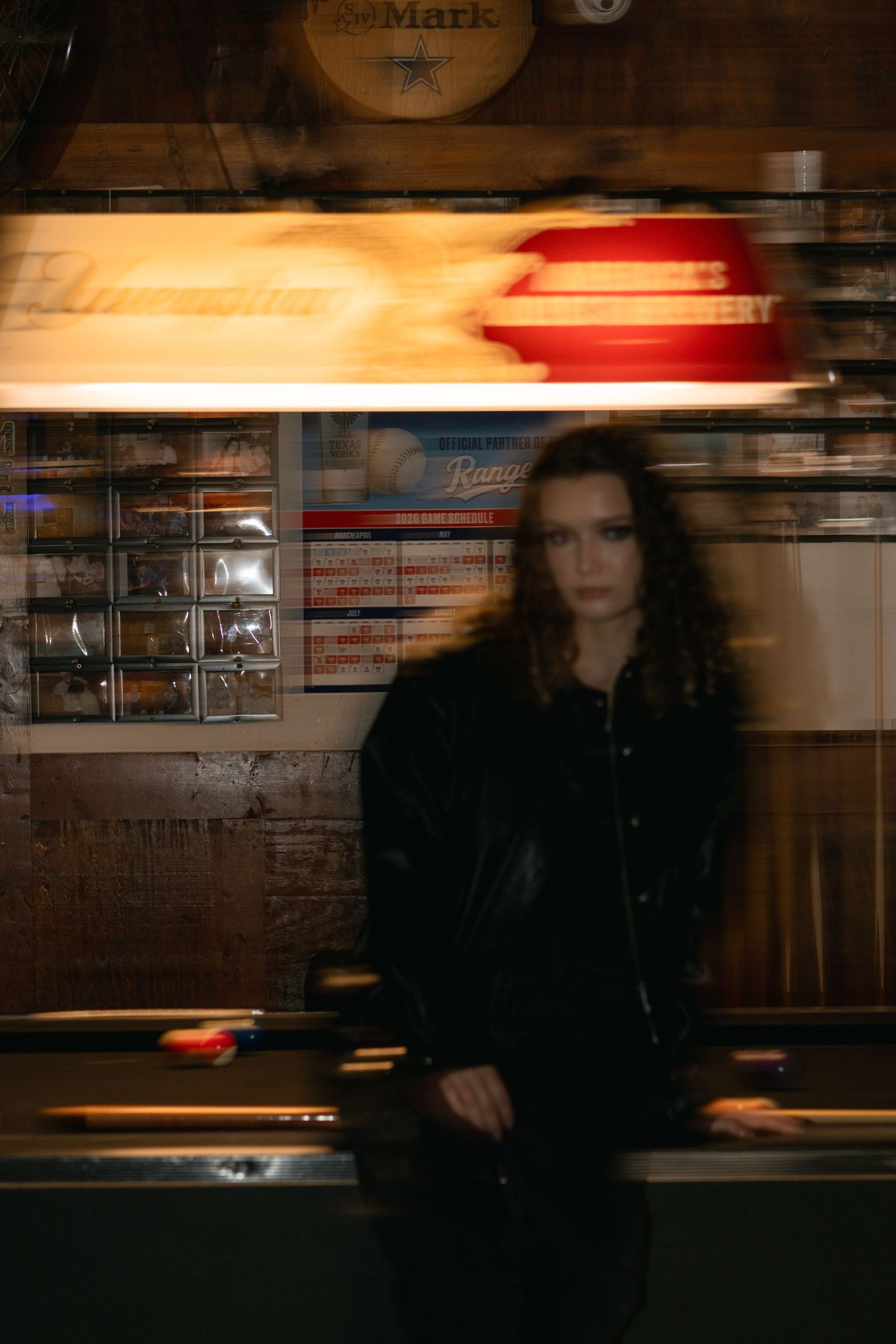 A young woman in a black leather jacket stands in front of a pool table in a dimly lit bar with motion blur effect.