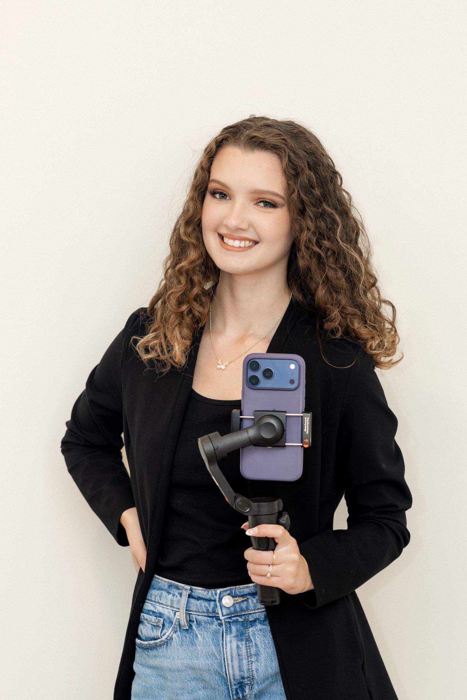 Smiling young woman with curly hair holding a smartphone gimbal stabilizer in black blazer.