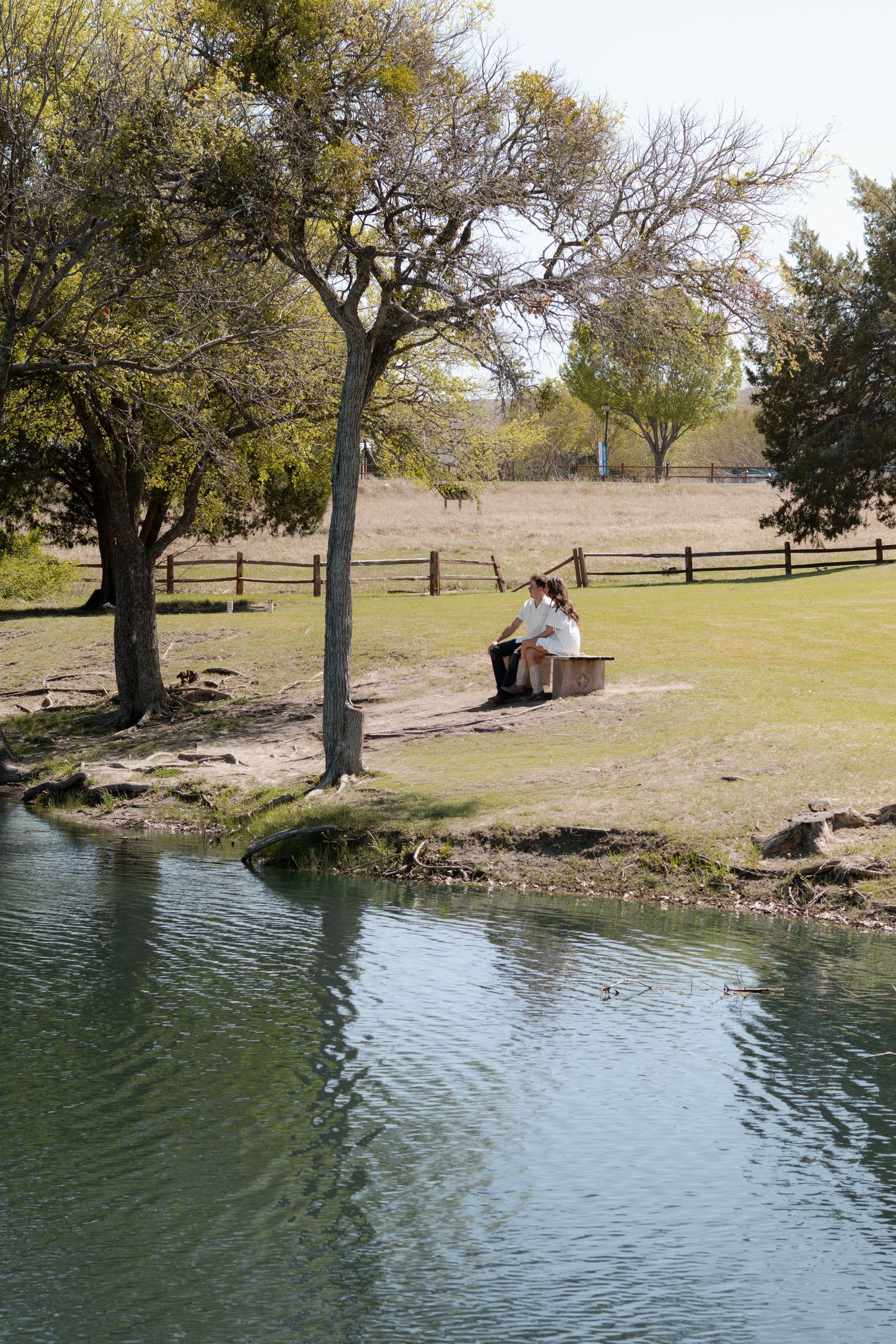 Person sitting on a bench by a tranquil pond in a lush green park with trees and wooden fence.
