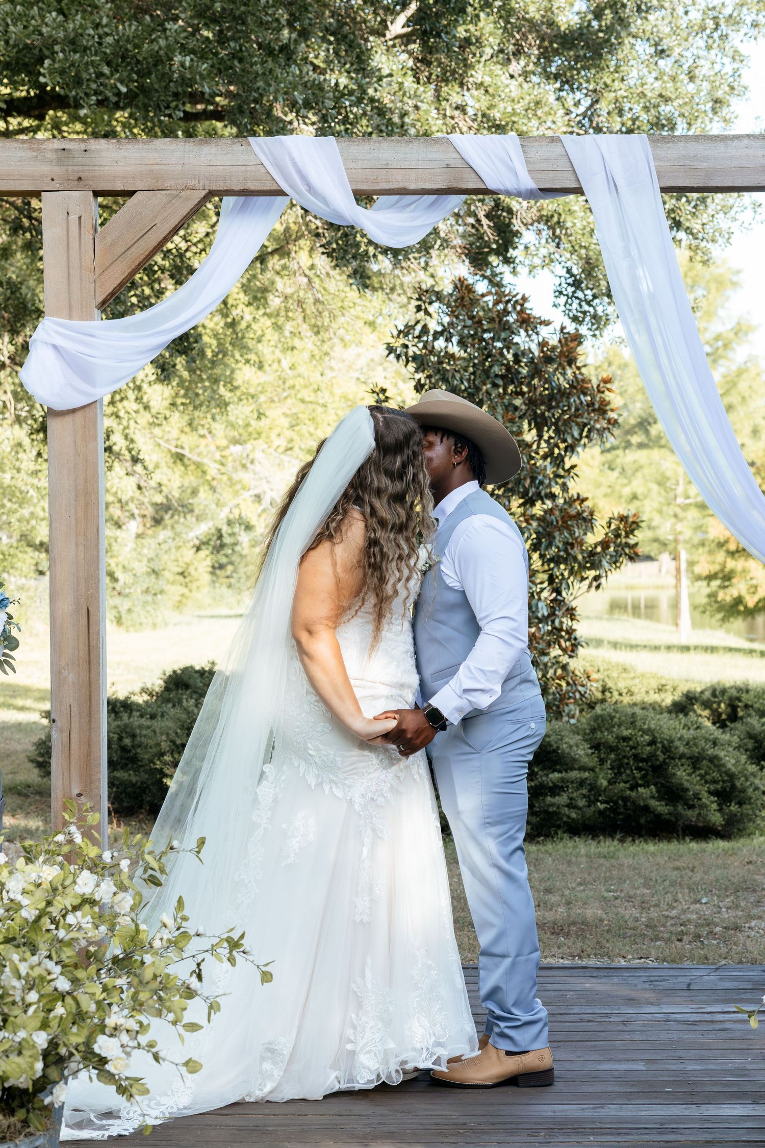 Bride and groom sharing a kiss under a rustic wooden arch with white draping.