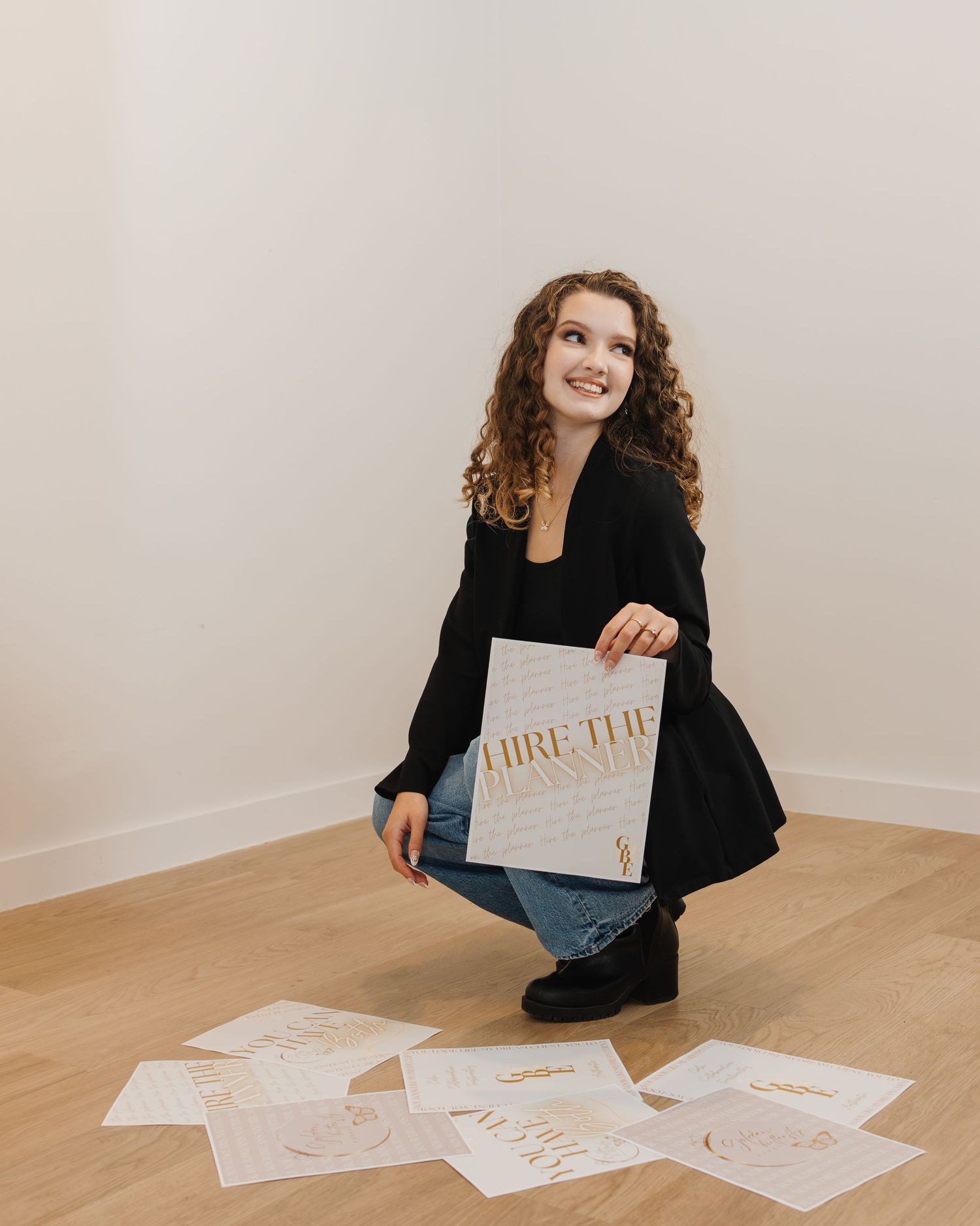Young woman with curly hair crouching on wood floor holding branding documents in black blazer and jeans.