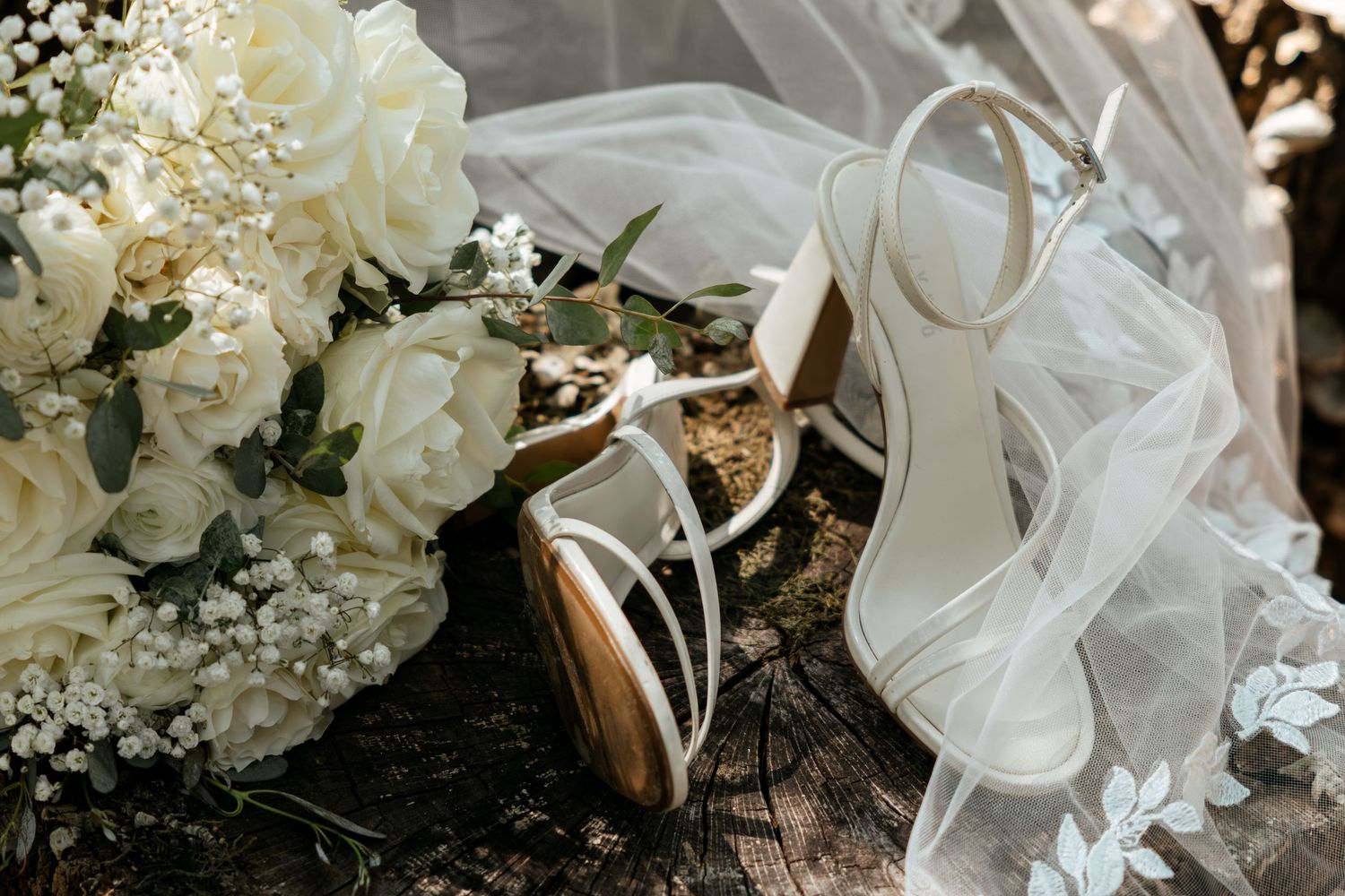White bridal heels rest beside a lush white rose bouquet and flowing lace veil on a rustic wooden surface.