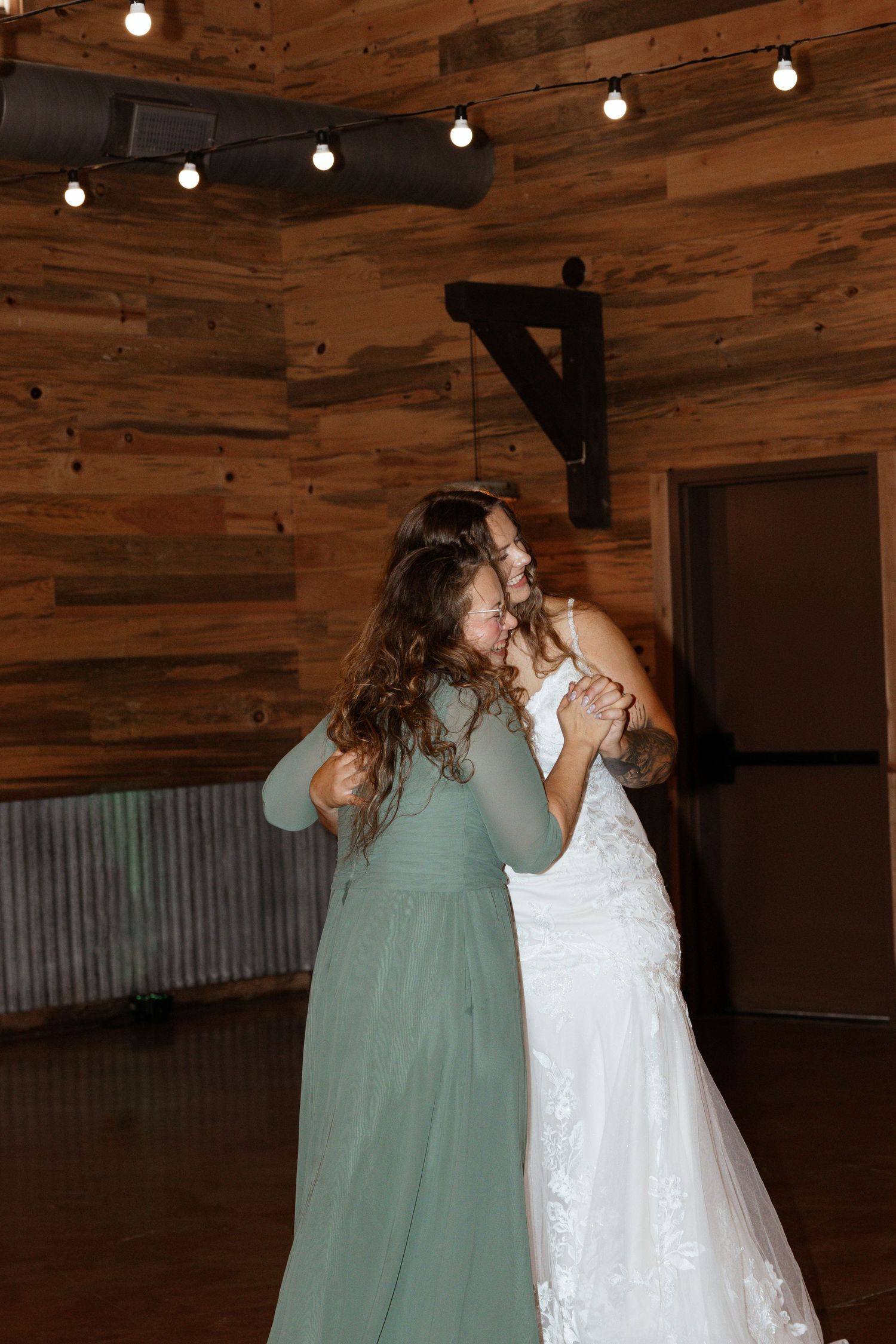Bride in white lace gown shares emotional dance with guest in rustic barn wedding venue.