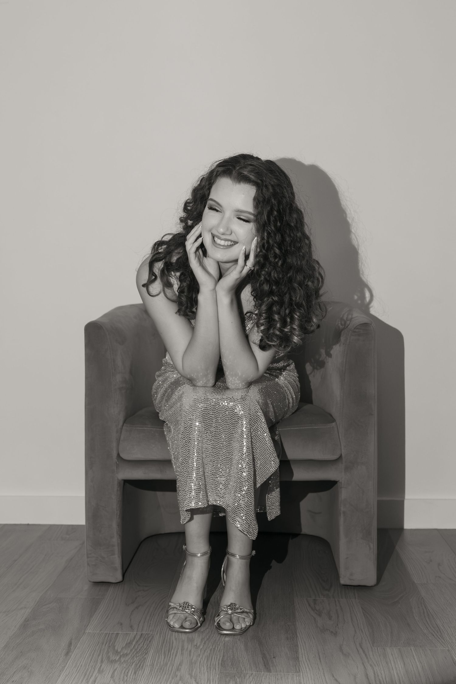 Black and white photo of a smiling young woman with curly hair sitting in a velvet chair.