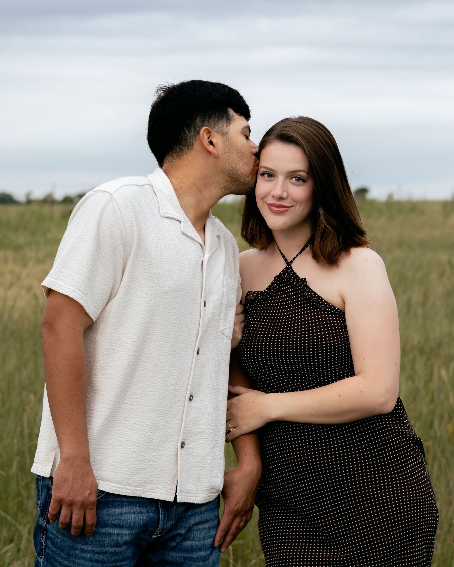 Couple in a green field, man kissing woman on cheek, she smiles in black polka dot dress.