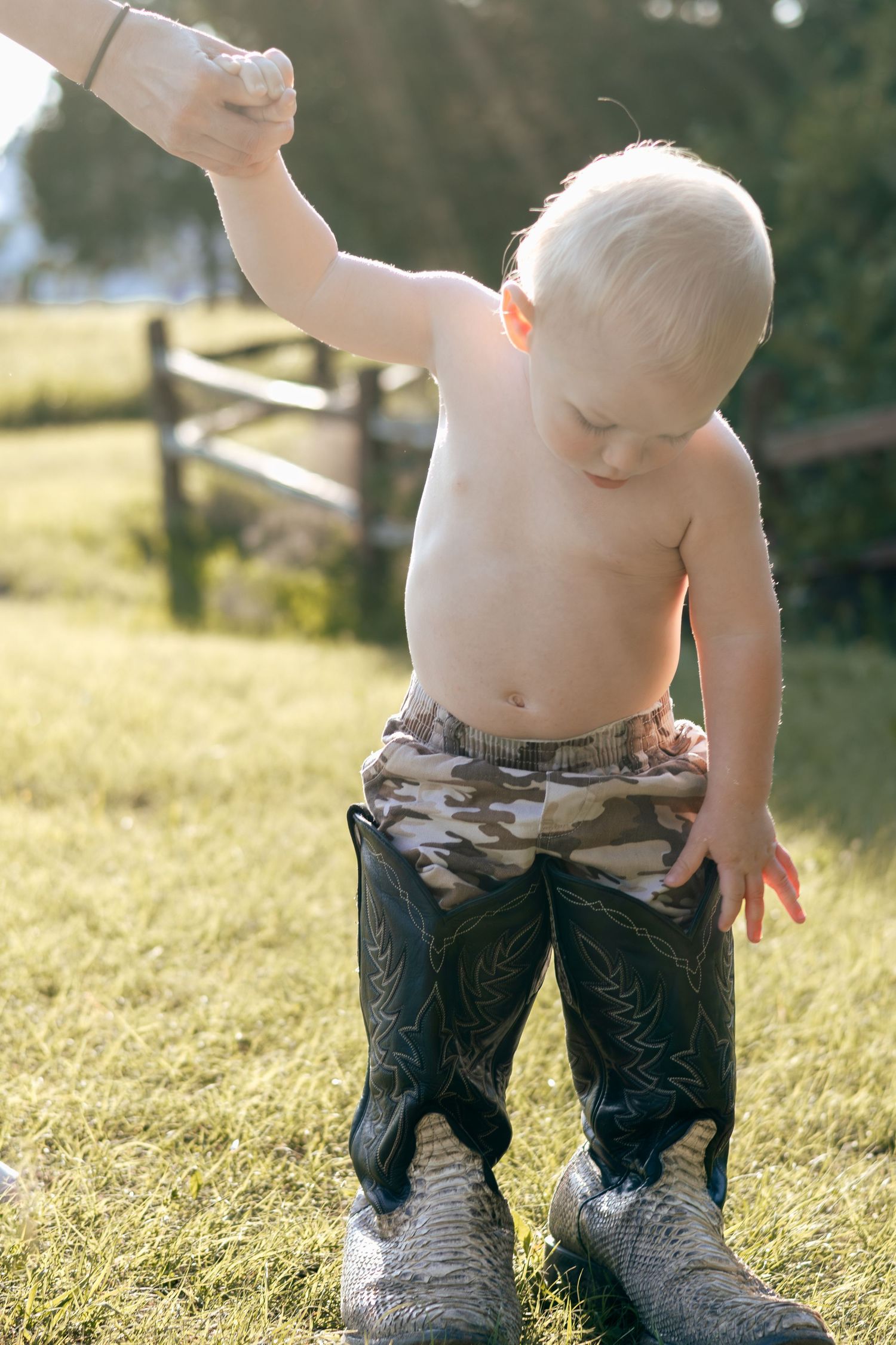 Shirtless toddler in camo shorts and oversized cowboy boots holds an adult hand while standing on green grass outdoors.