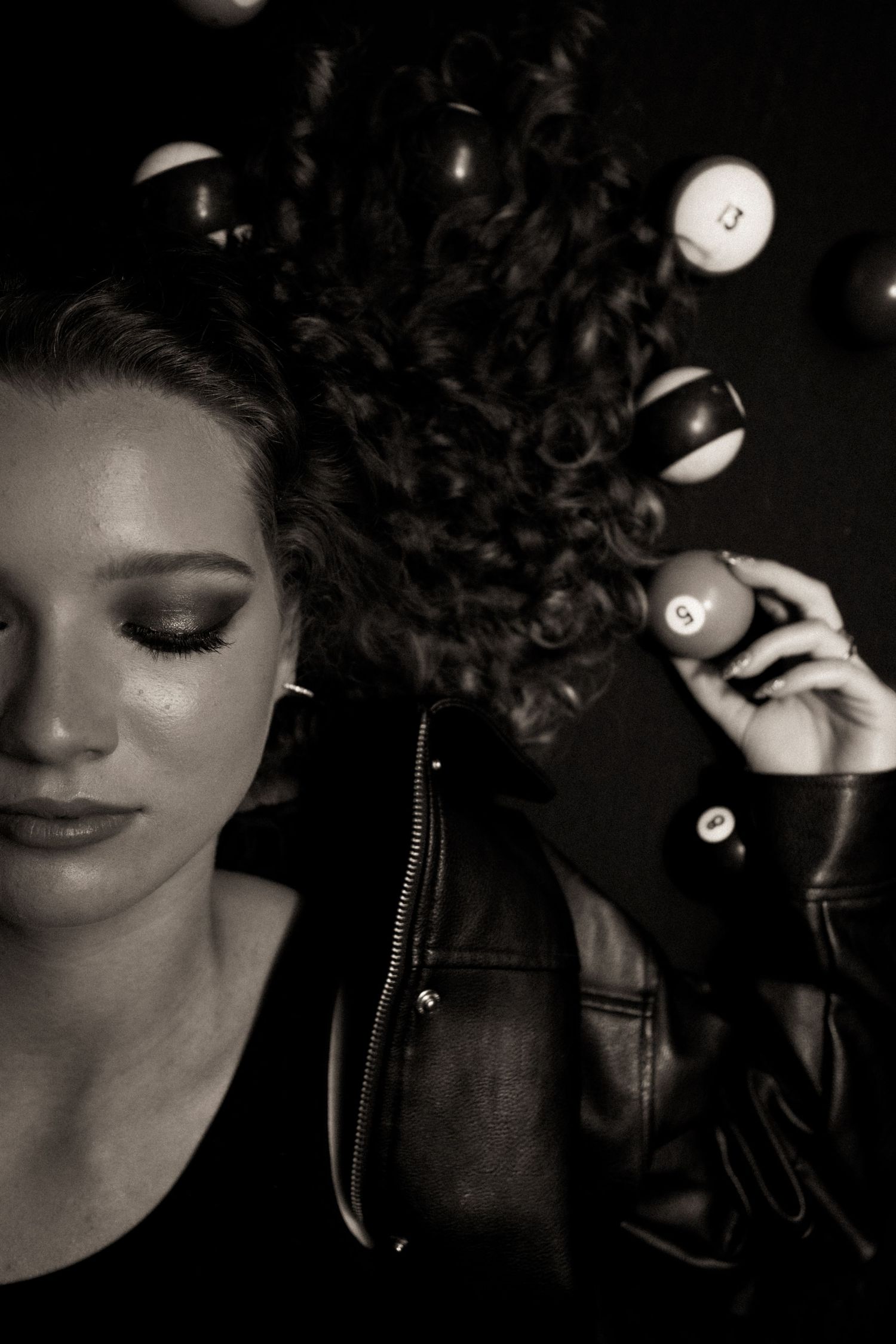 Black and white portrait of a woman in leather jacket surrounded by floating billiard balls with dramatic eye makeup.