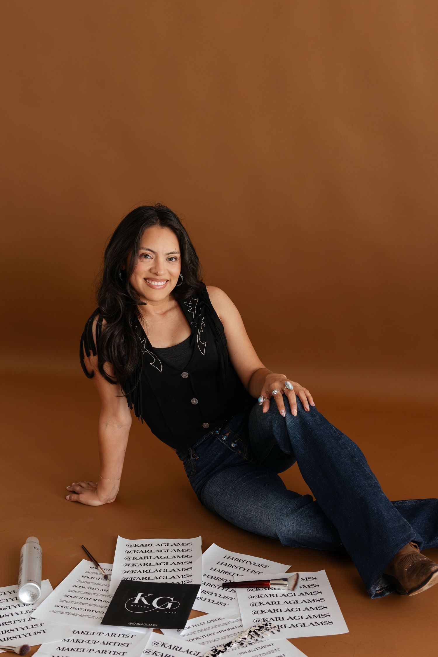 Woman in black vest and jeans sits on floor surrounded by KG makeup artist branded papers, smiling at camera.