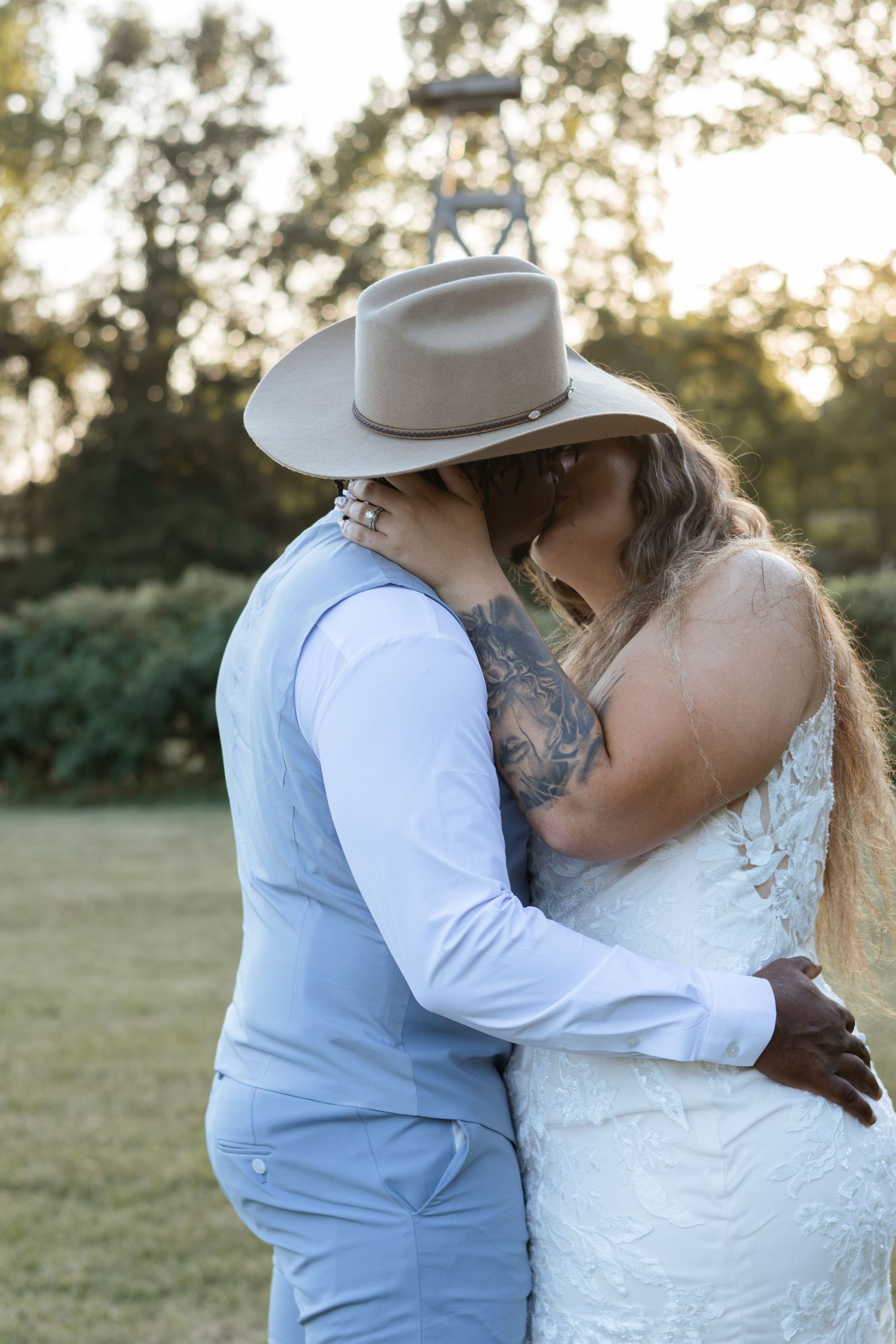 Newlyweds share their first kiss outdoors, groom in light blue suit and tan cowboy hat embracing tattooed bride.