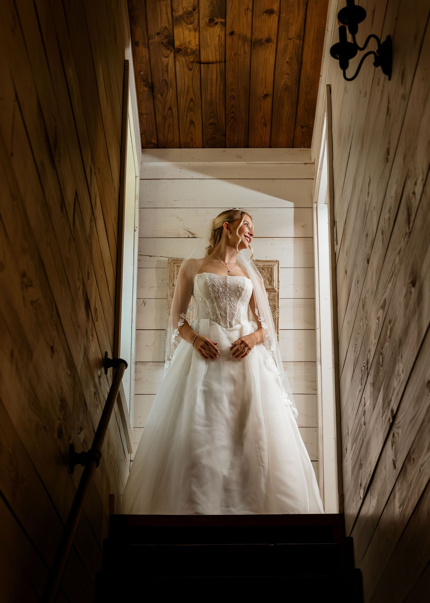 Bride in strapless lace ball gown wedding dress stands in rustic wooden stairwell bathed in natural light.