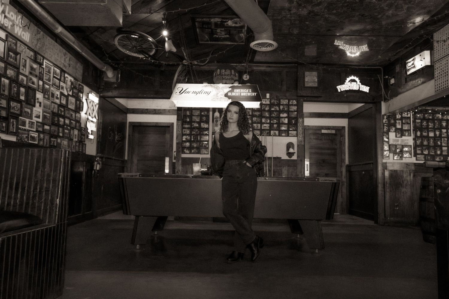 Woman in black outfit sits on a bench inside a vintage bar, captured in moody black and white photography.