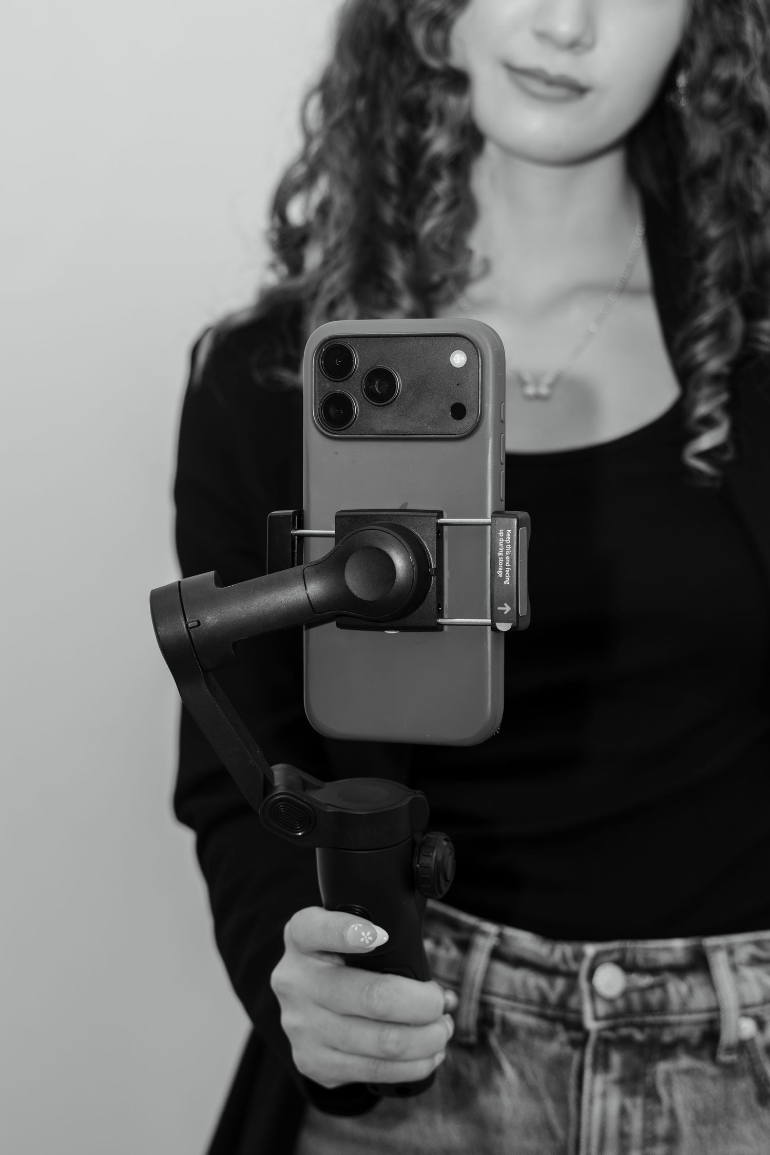 Young woman with curly hair holding a smartphone gimbal stabilizer, black and white portrait photo.